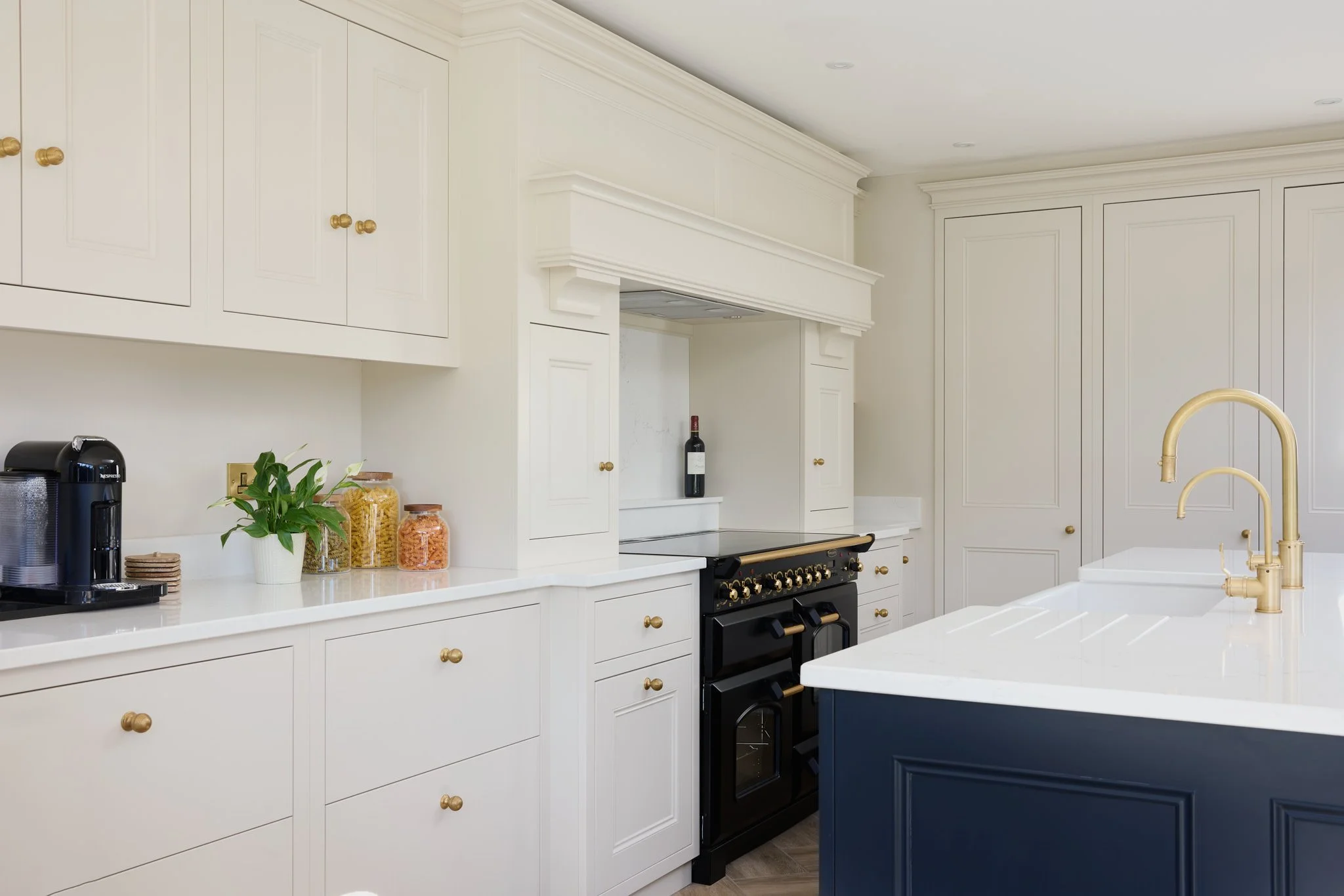 White kitchen with gold knobs, black stove, gold faucet, and a blue island, with cabinets, a wine bottle, and jars of pasta on the counter.