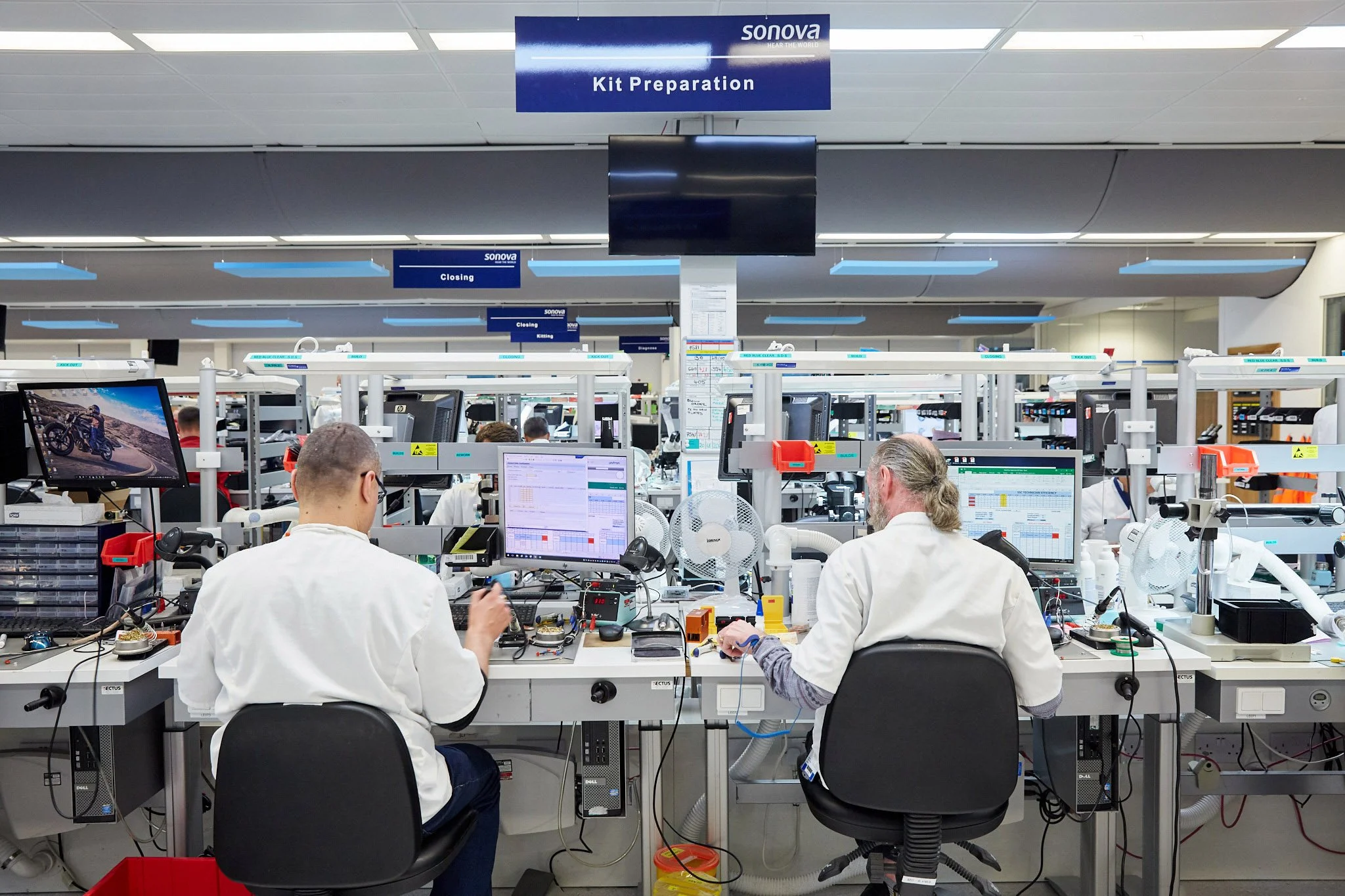 Laboratory workers in white lab coats working on electronic equipment at a bench with multiple computer monitors in a modern laboratory, under signs that read 'Kit Preparation' and 'Closing'.