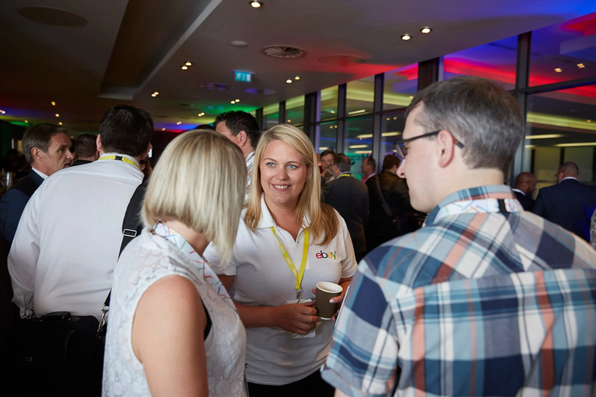 A group of people are socializing at a professional event or conference in a brightly lit room with colorful lights and large windows. A woman with blonde hair is holding a coffee cup and talking to others, wearing a white shirt with an eBay logo.