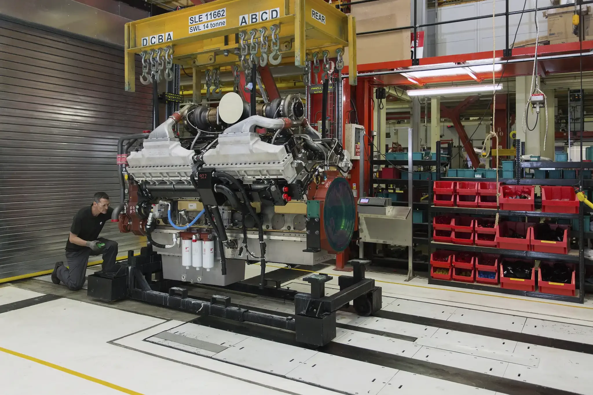 A worker in a factory wearing safety glasses, kneeling beside a large aircraft engine that is mounted on a black platform. The factory has shelves with red bins and industrial equipment, and overhead cranes with chains.