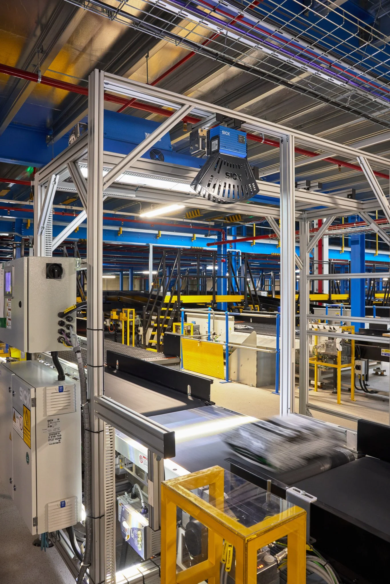 Industrial manufacturing plant with machinery, conveyor belt, and metal framing, including a large blue and black device and yellow safety barriers.