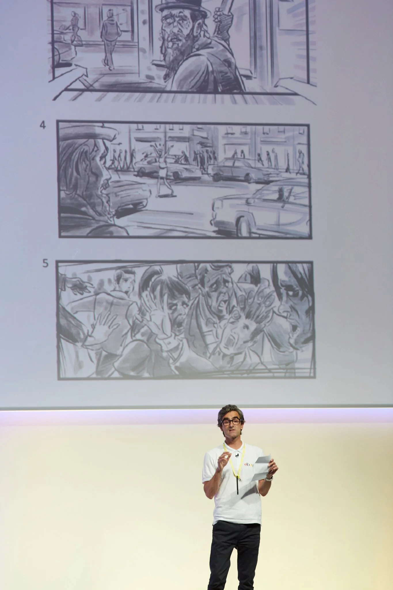 A man with glasses and dark hair, wearing a white eBay T-shirt, stands on stage holding a sheet of paper and a pen. Behind him is a large screen displaying a comic strip with three panels: the first panel shows a man with a beard and hat looking out 