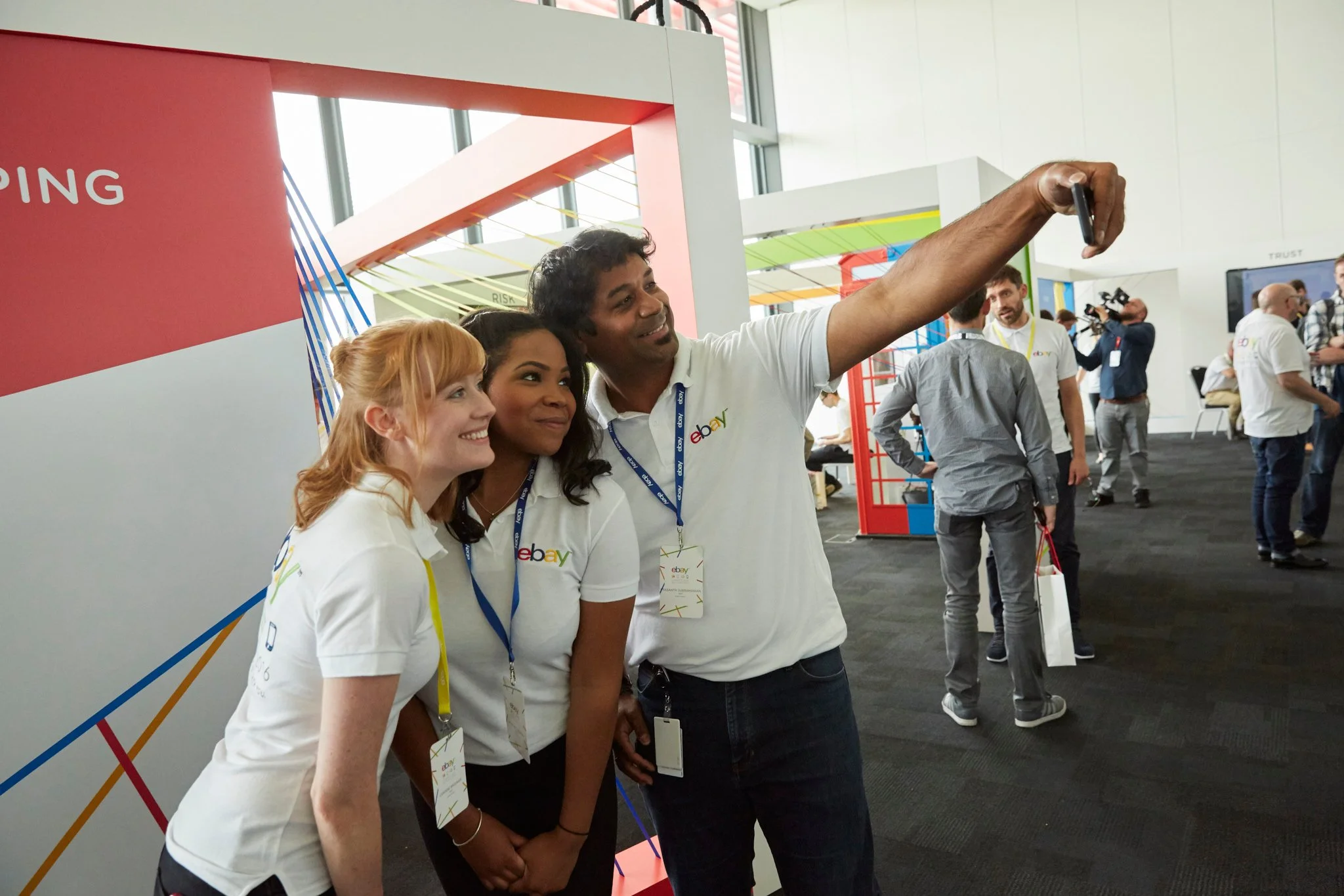 Three eBay employees taking a selfie at an event, with a man holding a smartphone extended to capture everyone in the frame. They are smiling and standing in front of a colorful exhibit booth with other attendees in the background.