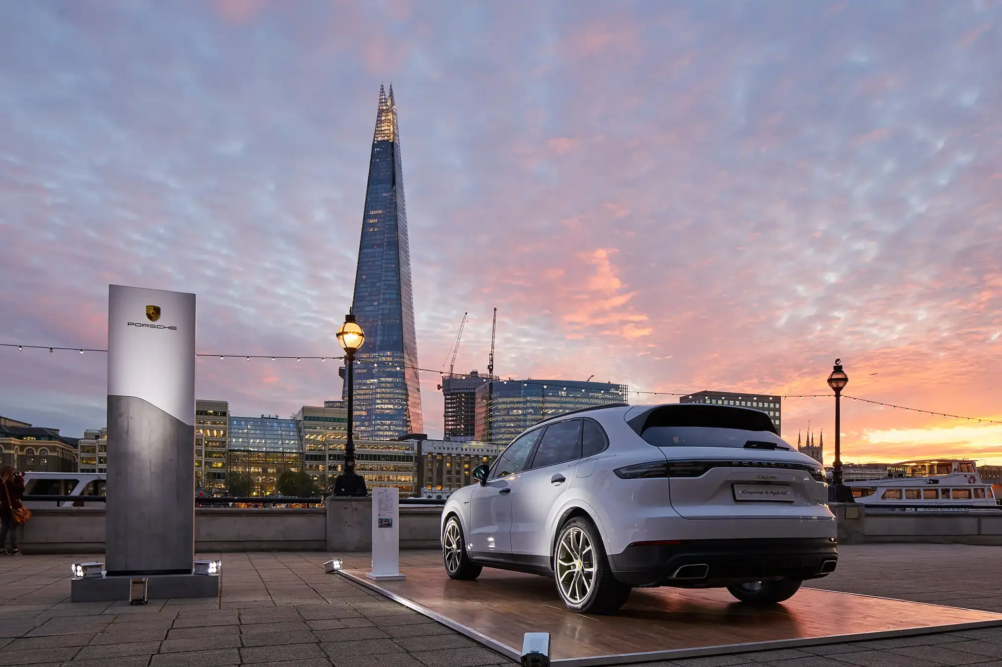 A white Porsche Cayenne e-Hybrid parked on a raised platform with a city skyline featuring the Shard skyscraper in the background during sunset. There are street lamps, a Porsche sign, and cranes in the cityscape.
