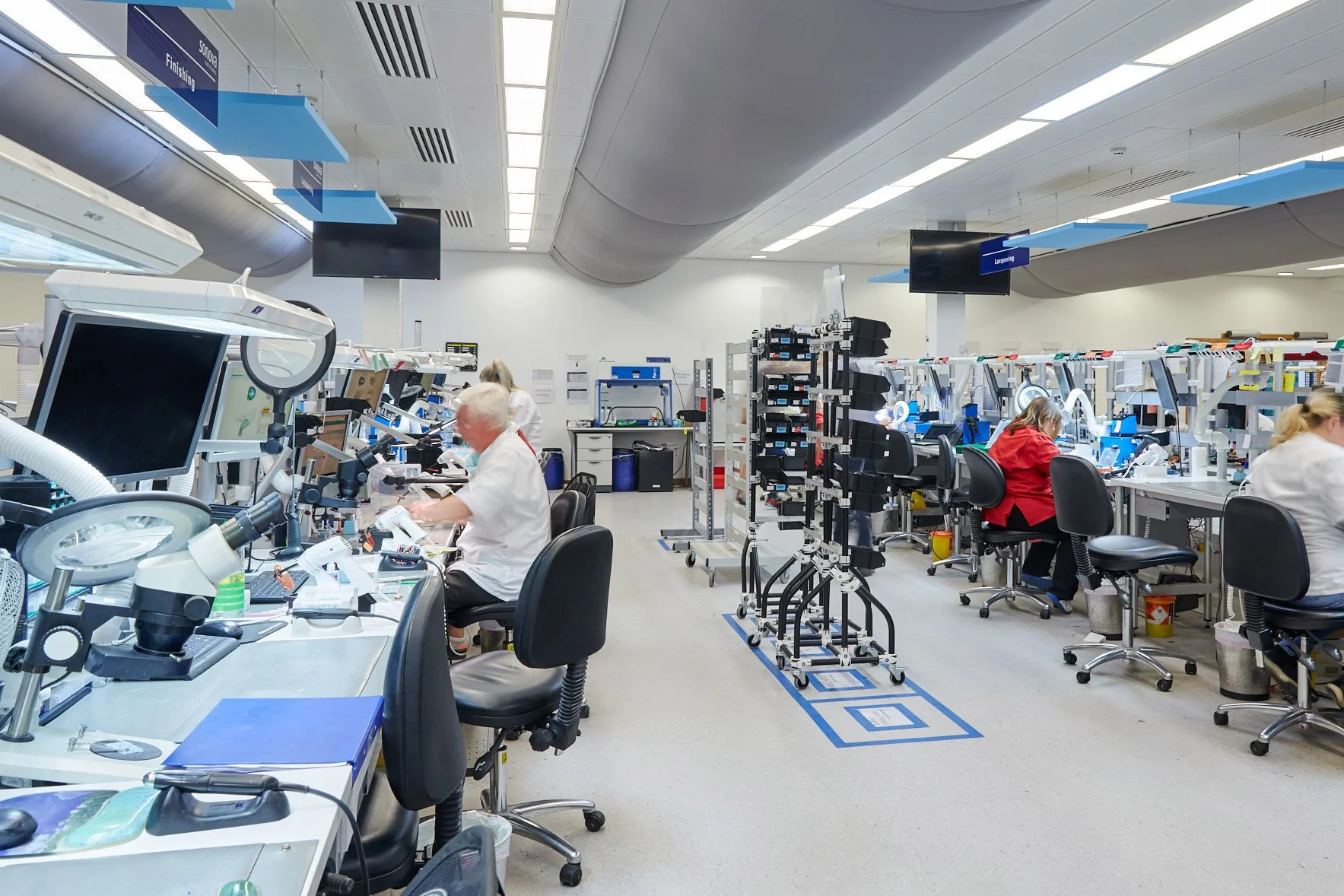 A clean, well-lit electronic manufacturing lab with workers at workstations involved in assembling electronic devices, along with equipment, tools, and monitors on desks, and overhead signs indicating different work stages.