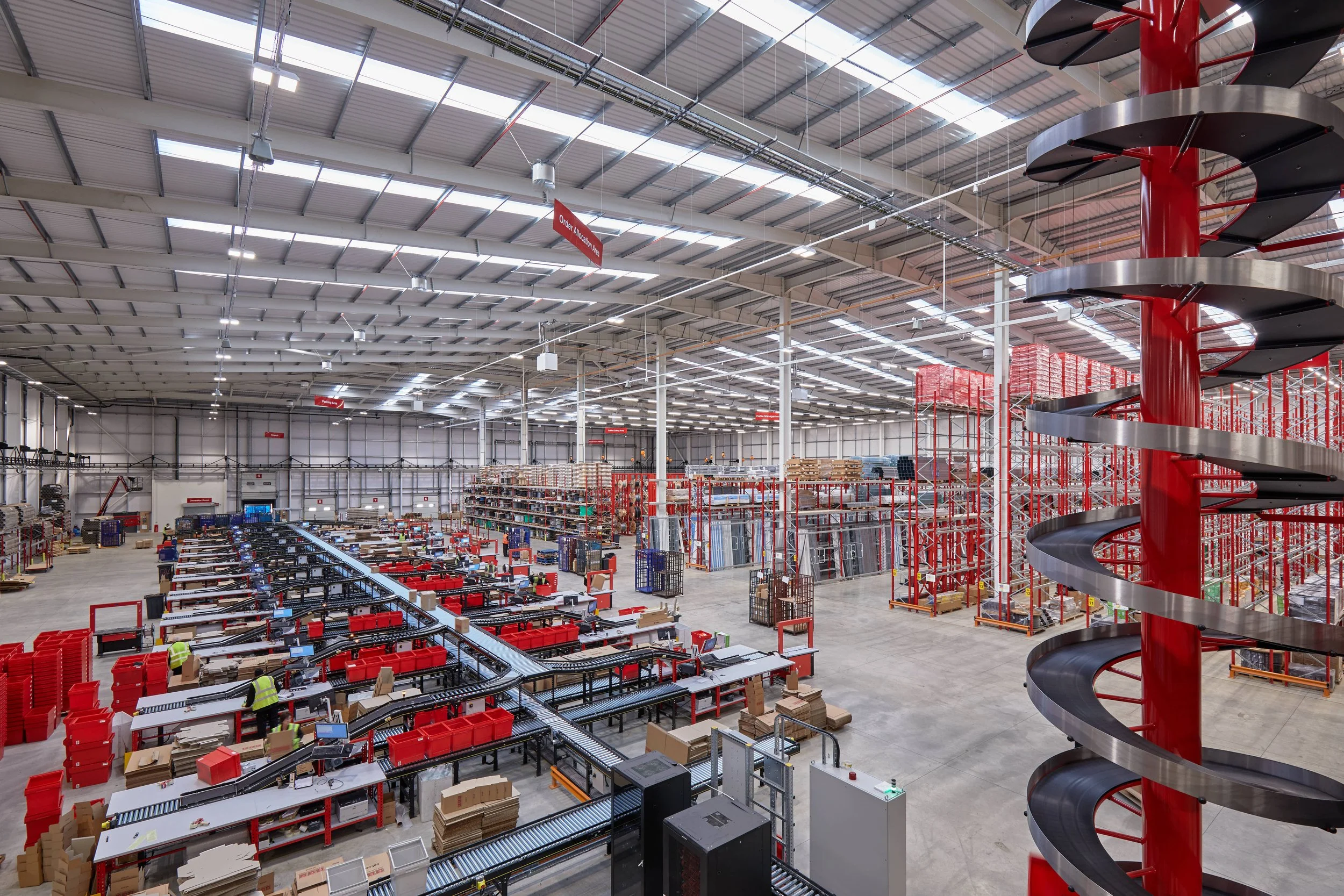Interior of a large warehouse or distribution center with high ceilings and metal roof. Red shelving units and conveyor belts are visible, with some workers present. A spiral slide with black steps and red sides is on the right side of the image. War