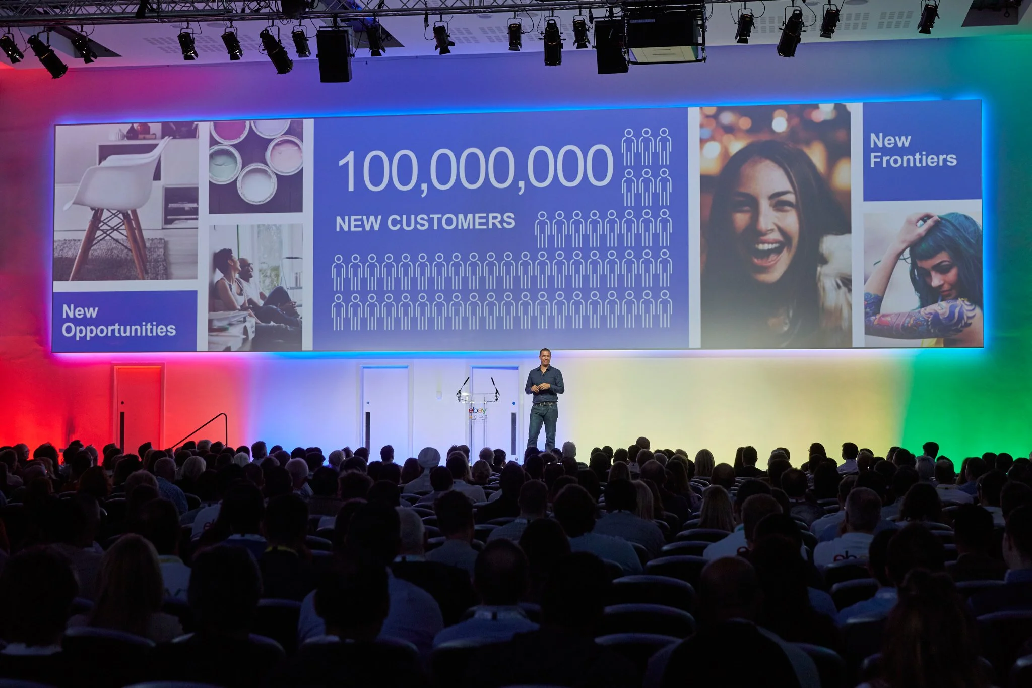 A speaker on a stage in front of a large screen displaying a presentation with statistics about new customers and opportunities, with a large audience seated facing the stage in a conference hall with colorful lighting.
