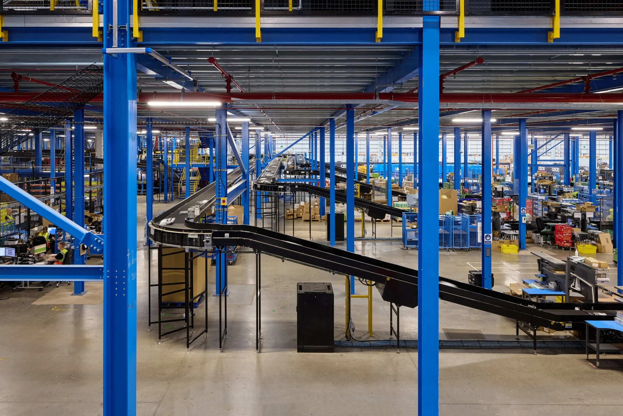Interior of a large warehouse with blue structural columns, conveyor belts, and storage areas filled with boxes and packages.