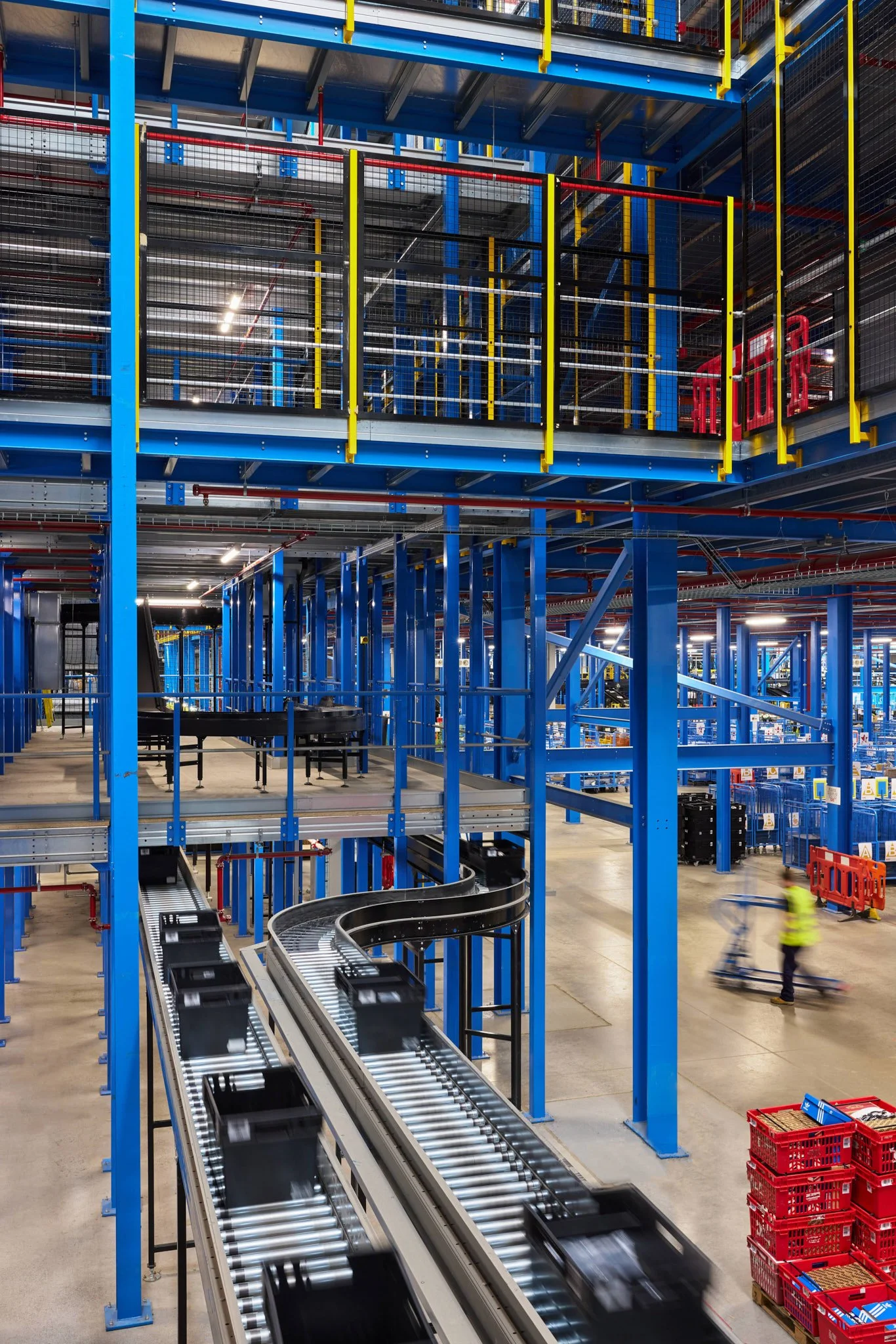 Inside a warehouse with multiple blue metal shelving racks, conveyor belts, and red crates. Two workers in safety vests are visible on the right side, moving crates.