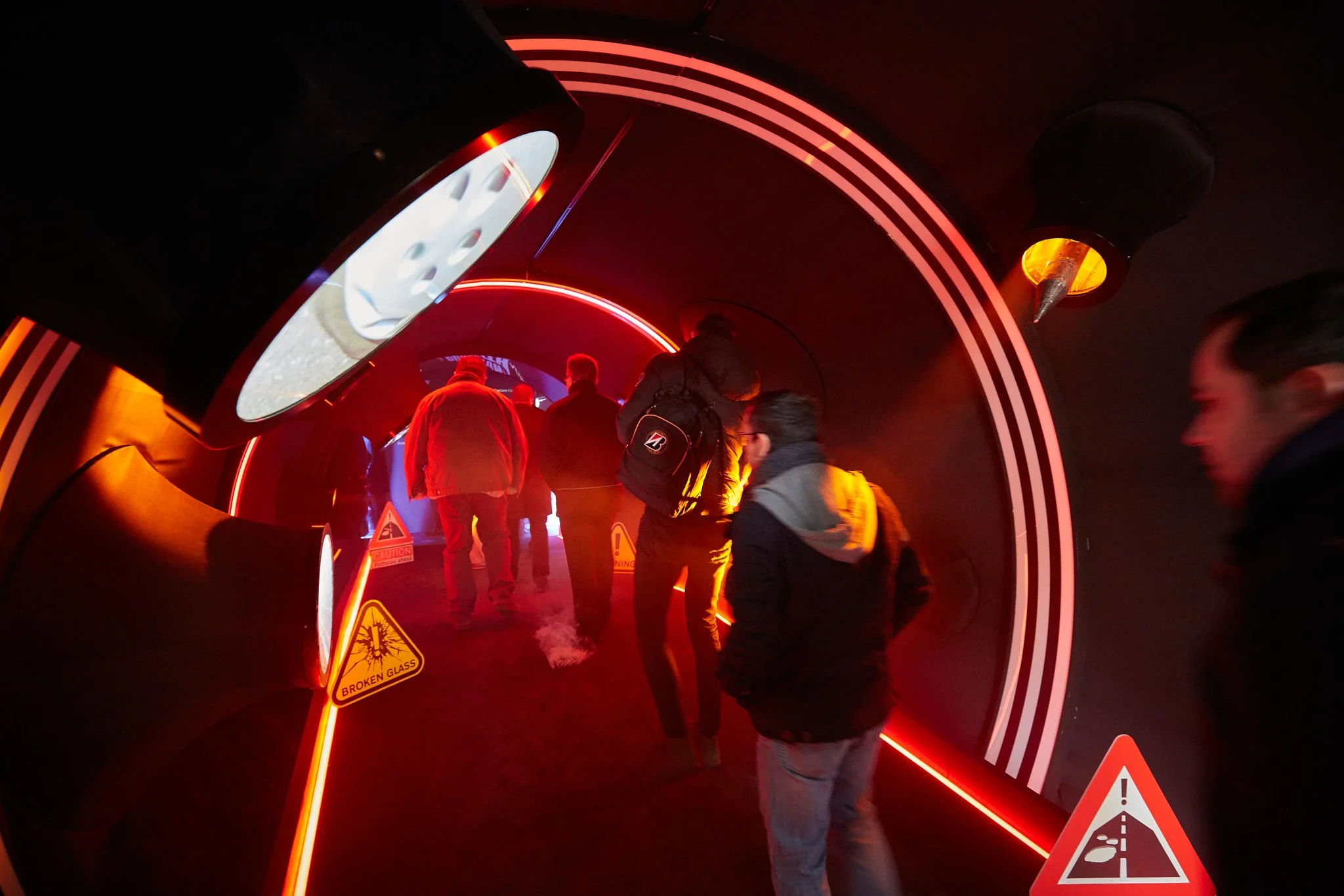 People walking through a tunnel with neon red and orange lights, warning signs about broken glass and a slip hazard, and illuminated warning signs on the ground.