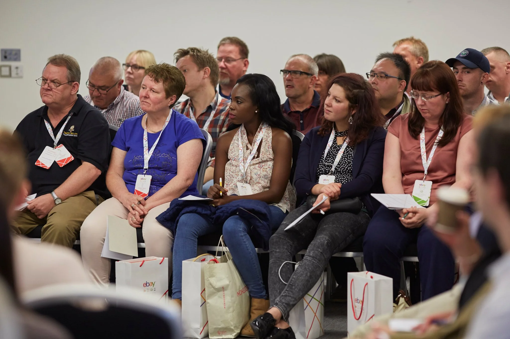 Conference attendees seated in rows listening to a speaker, with some holding notepads and wearing badges, and shopping bags on the floor in front of them.