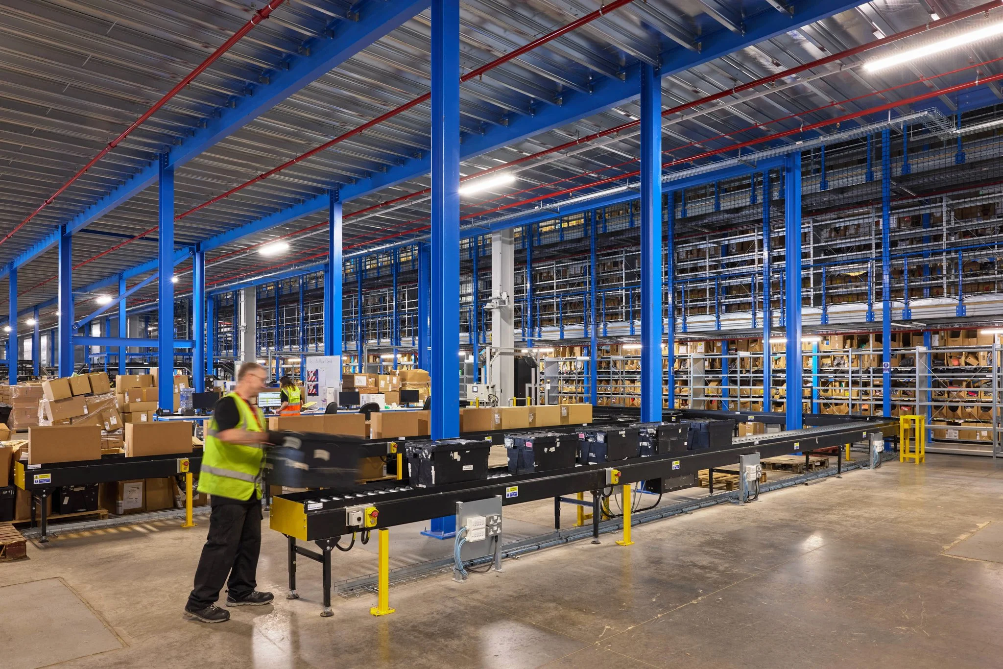 Inside of a large warehouse with blue shelving units filled with boxes and items. Workers in high-visibility vests operate conveyor belts and other machinery for sorting and moving packages.