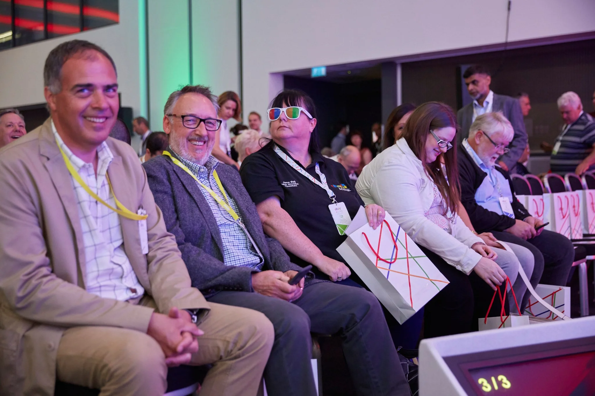 People attending a conference or seminar, seated in a row, with some holding gift bags, and one person wearing 3D glasses.