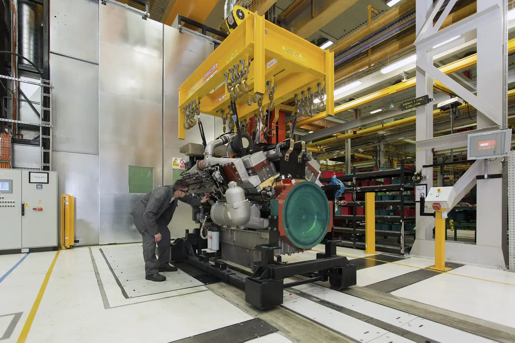 A scientist or engineer in a gray suit and glasses is working with a large piece of machinery in an industrial lab or manufacturing facility. The machine has a large green circular component and various pipes and parts. There are yellow safety barrie