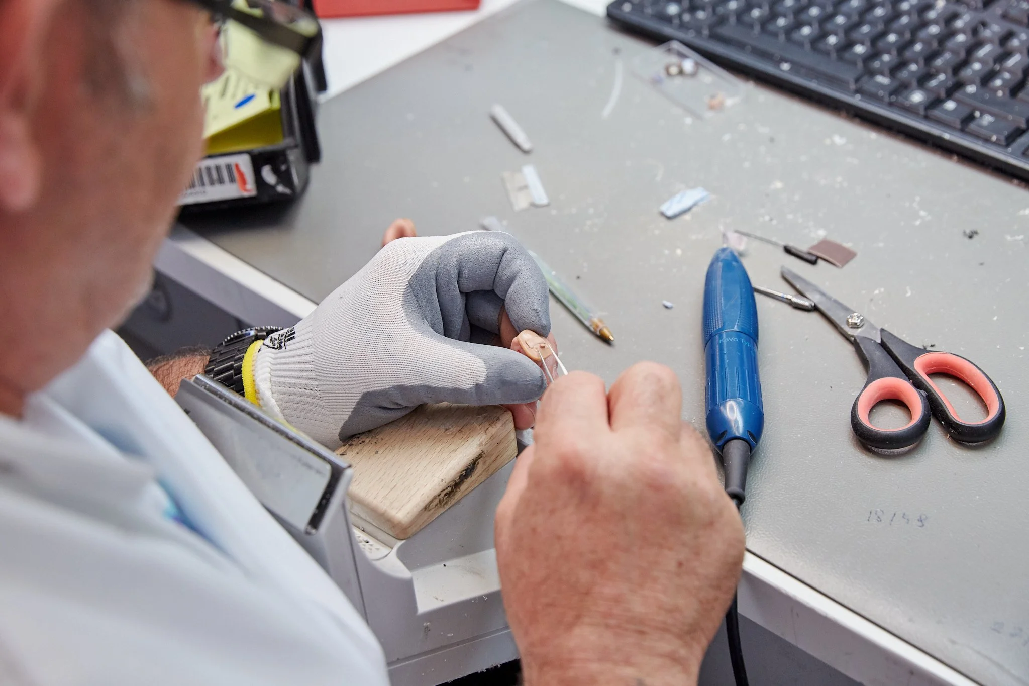 A person wearing a white glove and glasses working on a small electronic device at a cluttered workspace with tools, scissors, a blue pen, and a keyboard.