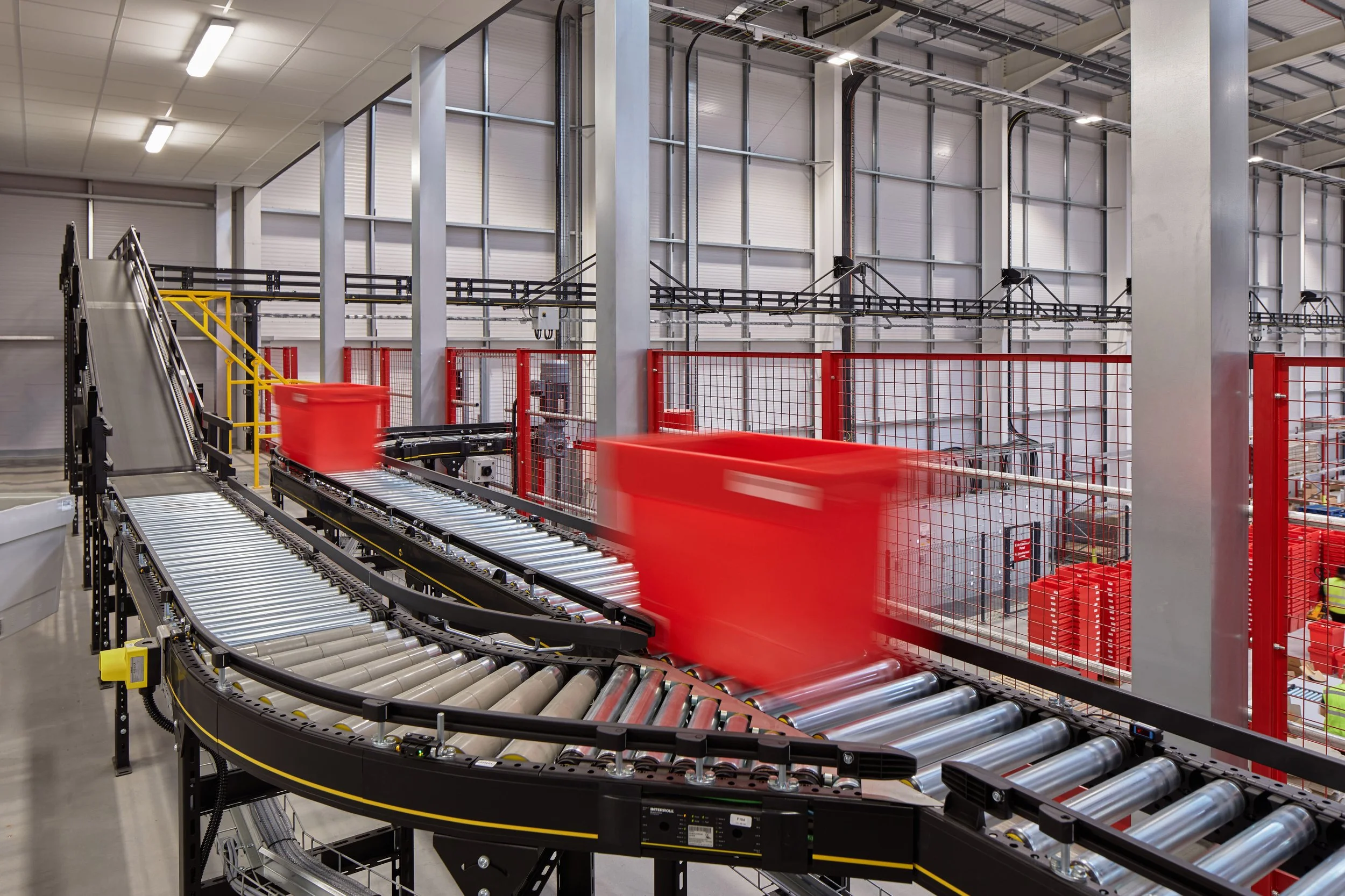 An automated conveyor system inside a warehouse, with red bins moving along a metal track and a red safety fence on the side.