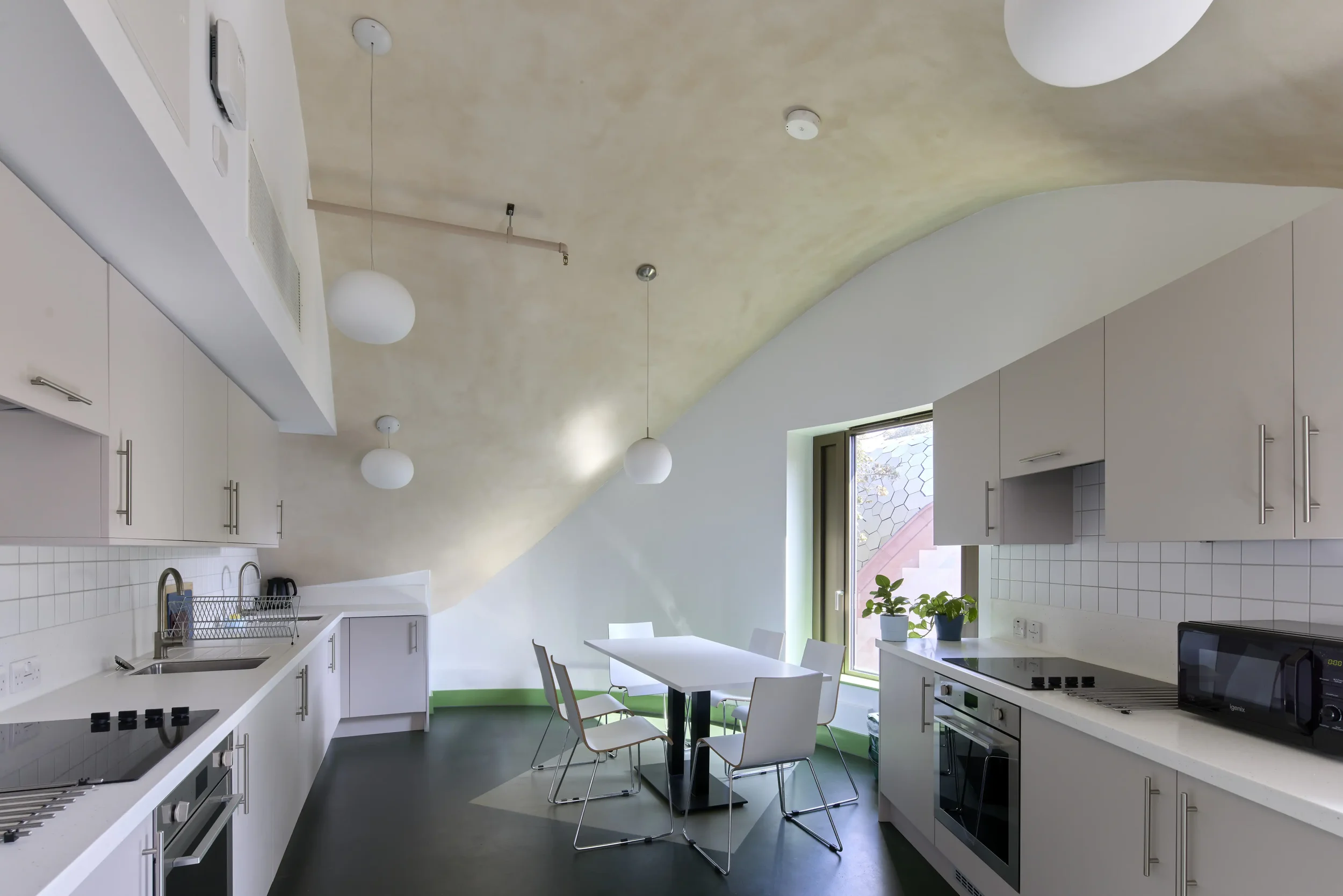Modern kitchen with white cabinets, a white countertop with a sink, a stovetop, microwave, and oven. A dining table with four chairs is in the center, and there are large windows with natural light. The ceiling has a curved design with hanging spheri