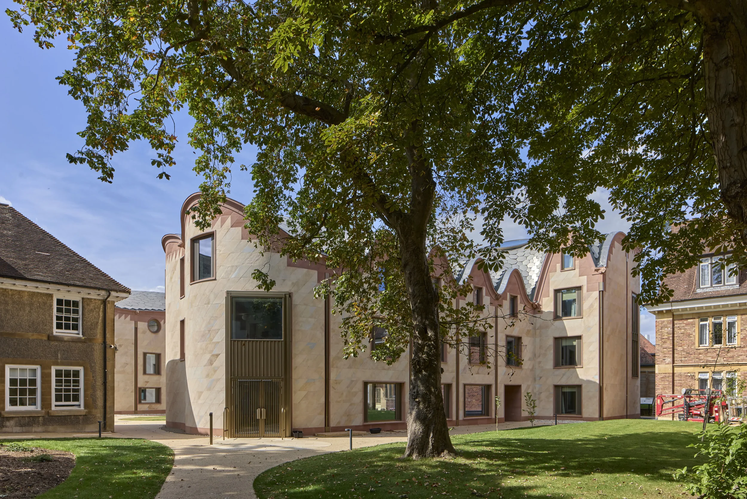 A modern residential building with a curved design, beige stone facade, and multiple windows, surrounded by a landscaped yard with green grass, a walking path, and a large tree with green leaves.