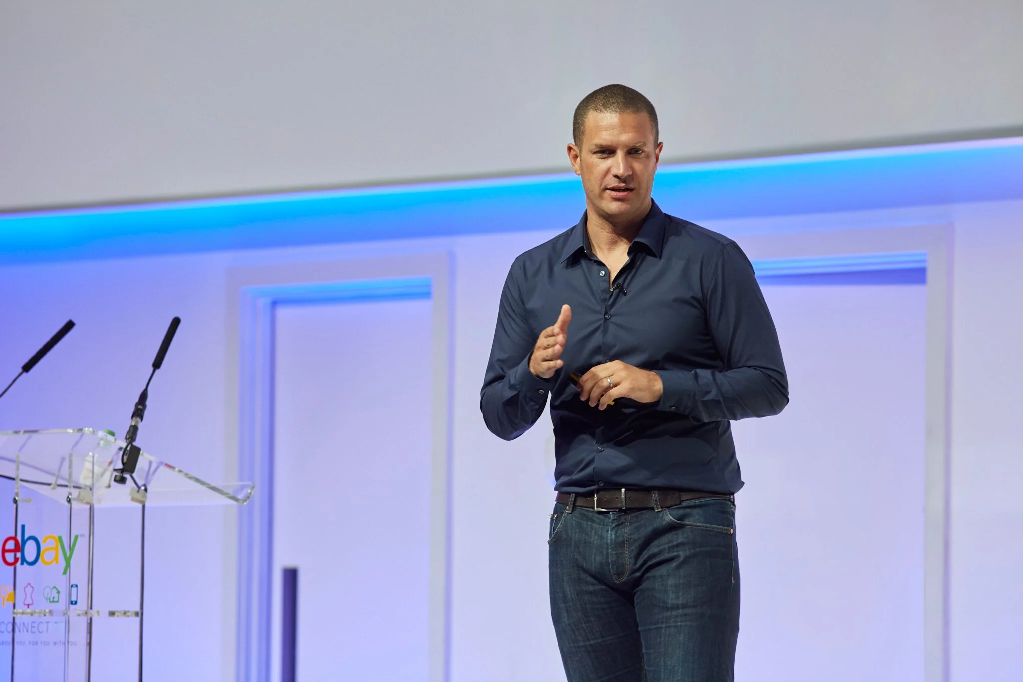 A man with short hair wearing a dark blue button-up shirt and jeans stands on stage during a presentation at eBay.