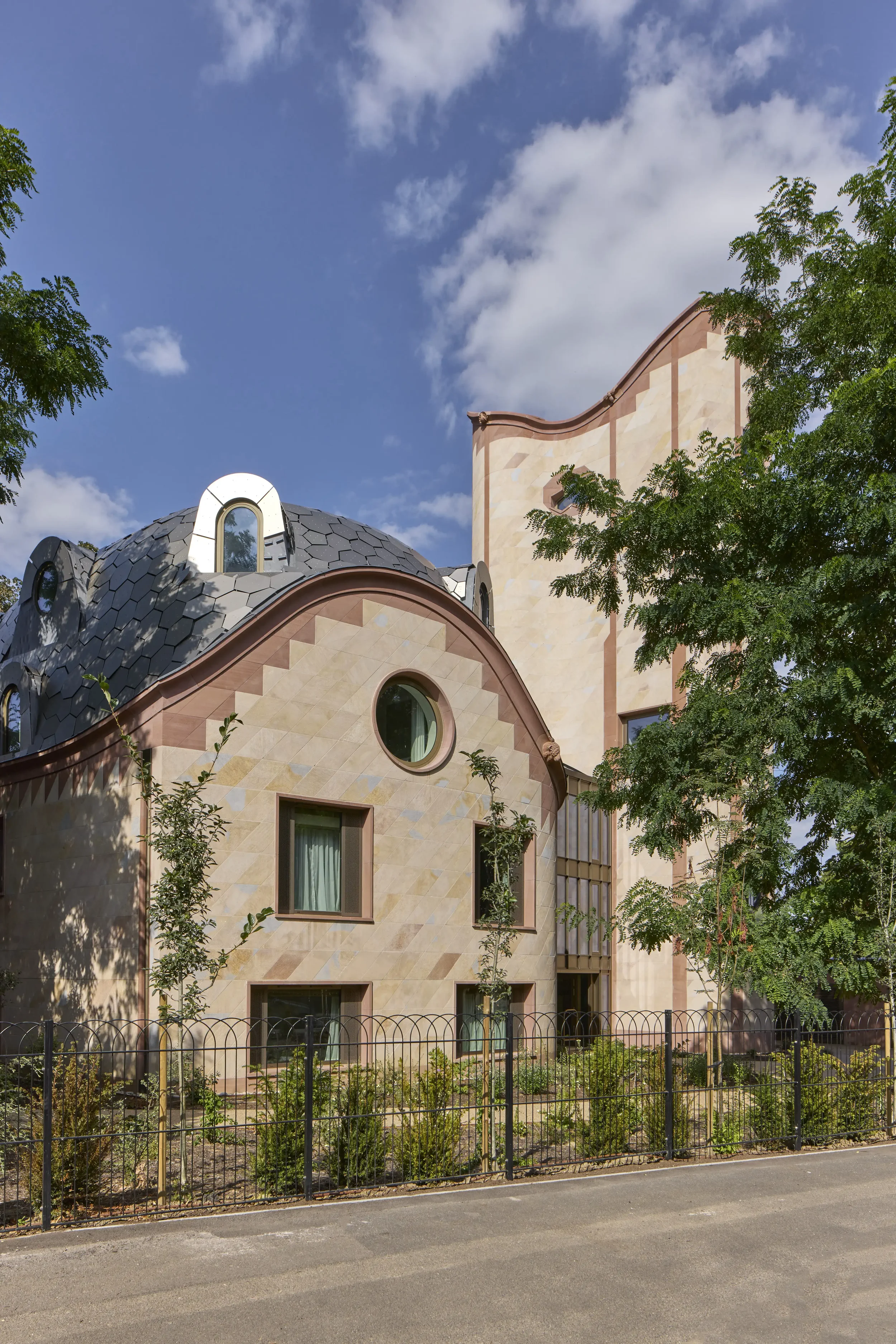 A modern building with rounded and curved architectural design, beige and pinkish exterior walls, multiple windows, and a slate shingle roof, surrounded by green trees and a black metal fence, under a partly cloudy blue sky.