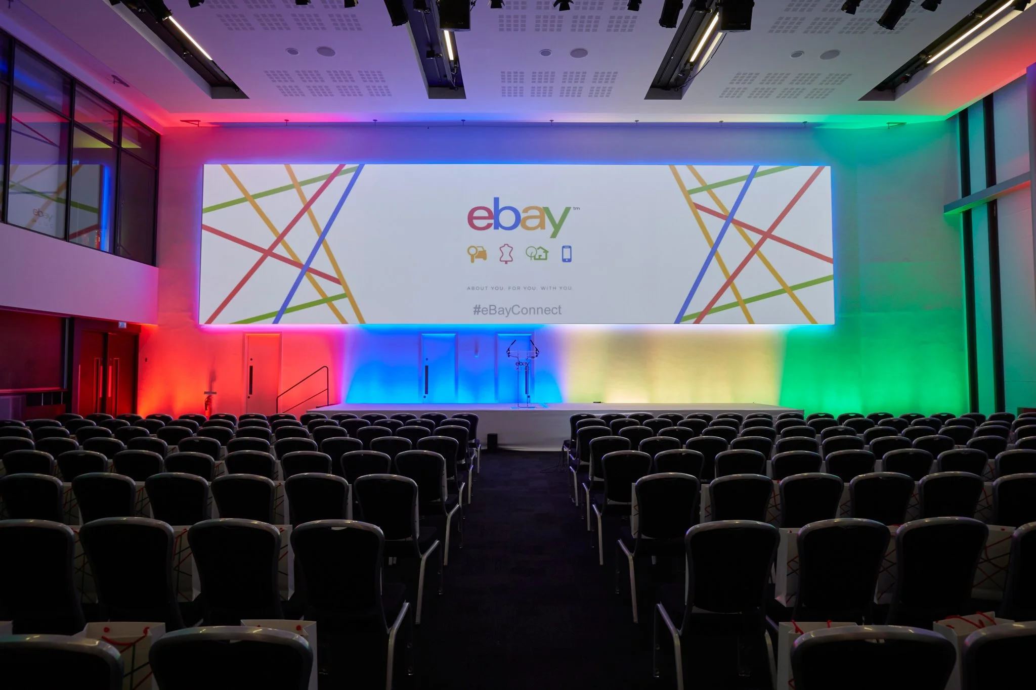 An empty conference room with a large screen displaying the eBay logo, colorful overlay lighting, and rows of chairs facing the stage.