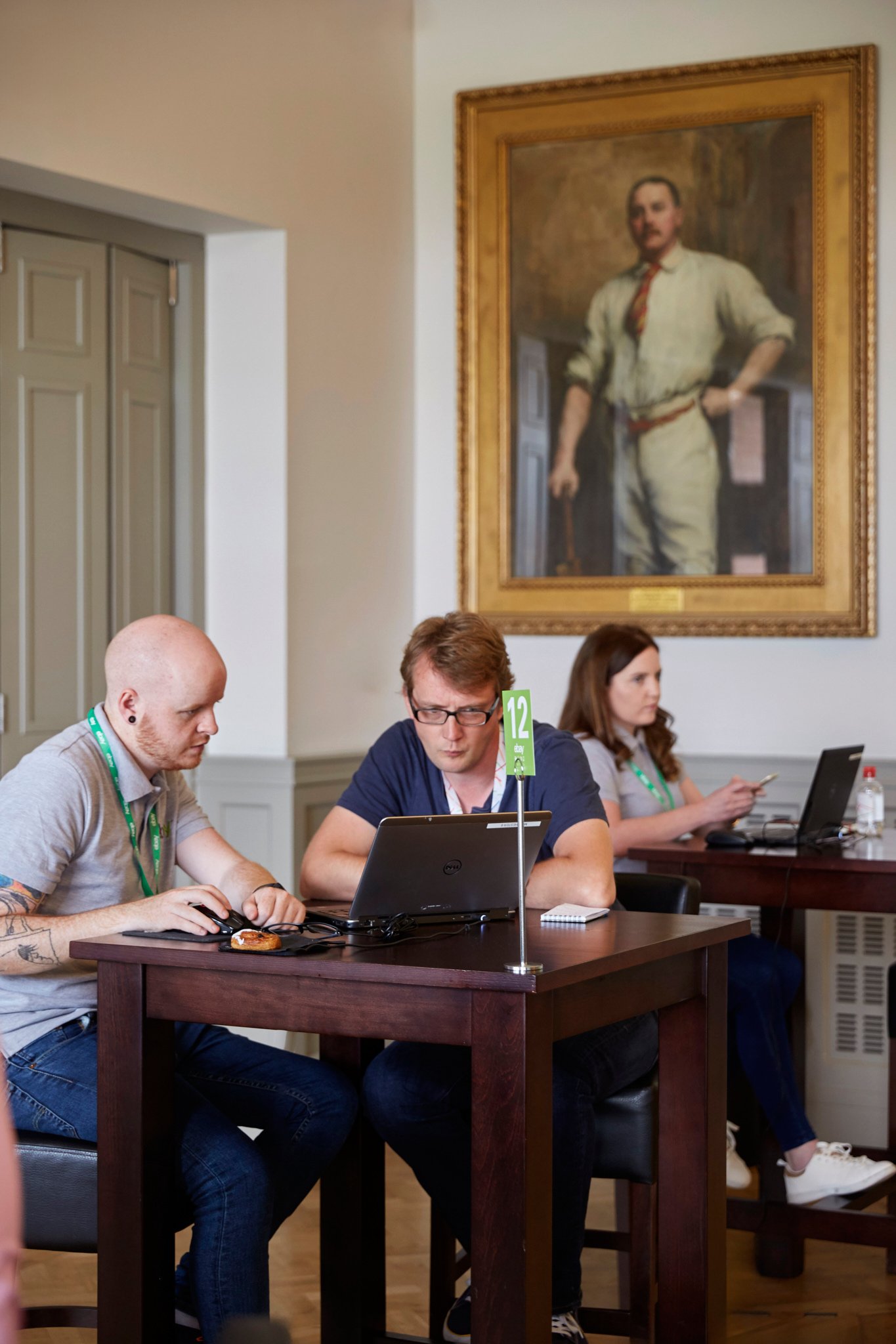 Three people sitting at a wooden table working on laptops in a room with a large portrait painting of a man in vintage clothing hanging on the wall behind them.