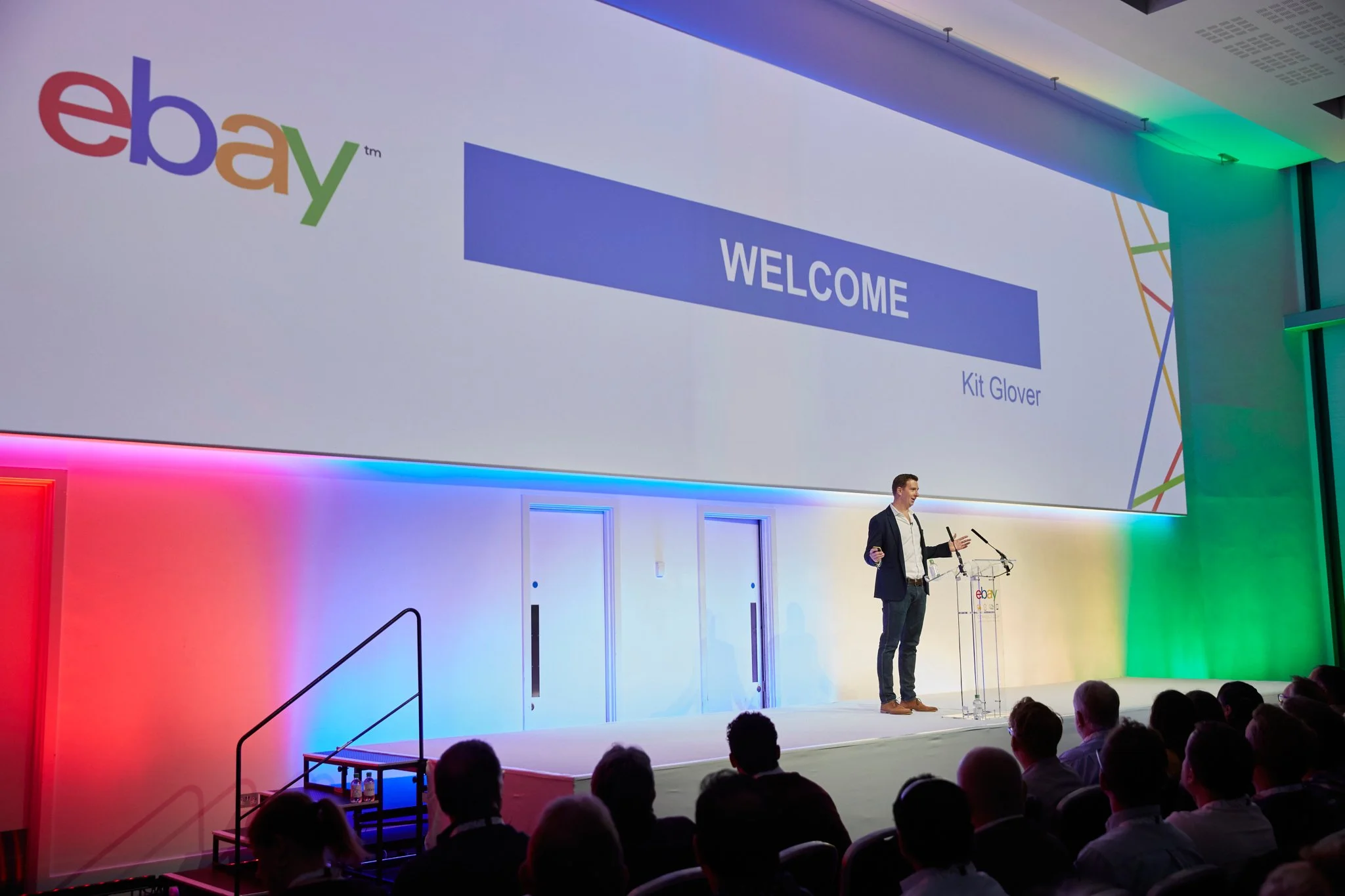A man gives a presentation on stage at an eBay event, with a large screen behind him displaying the eBay logo and a welcome message for Kit Glover. The stage is lit with colorful lighting.