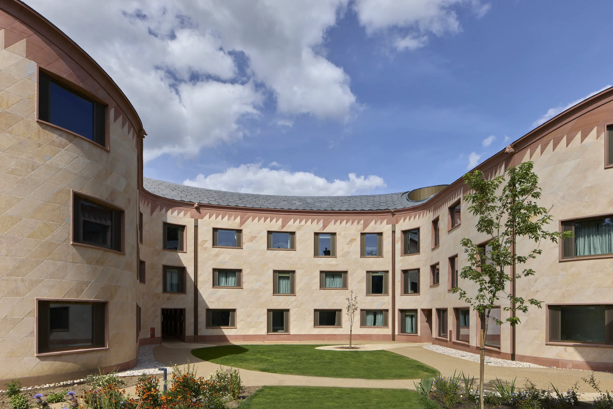 Curved beige building with multiple windows and a tiled roof, surrounded by a garden with trees and flowers under a blue sky with clouds.