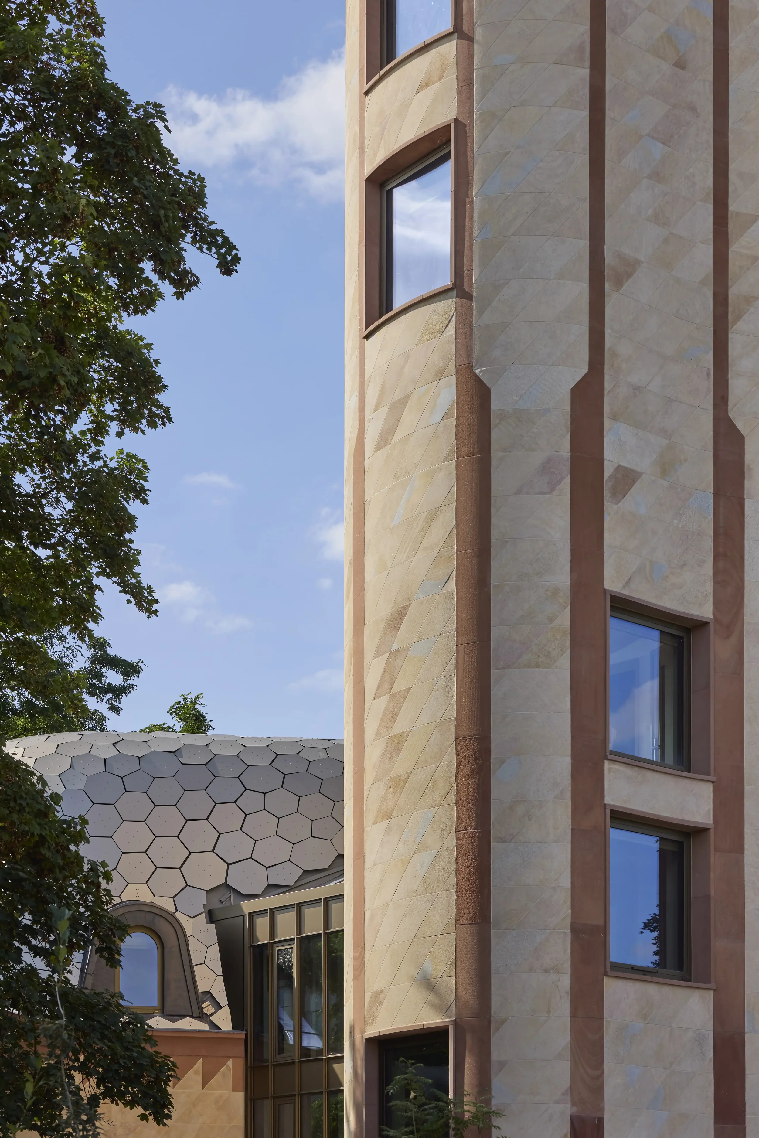 Close-up of building facades with unique architectural design, one with rounded windows and beige stone exterior, and another with geometric patterned roof and arched windows, surrounded by trees against a blue sky.
