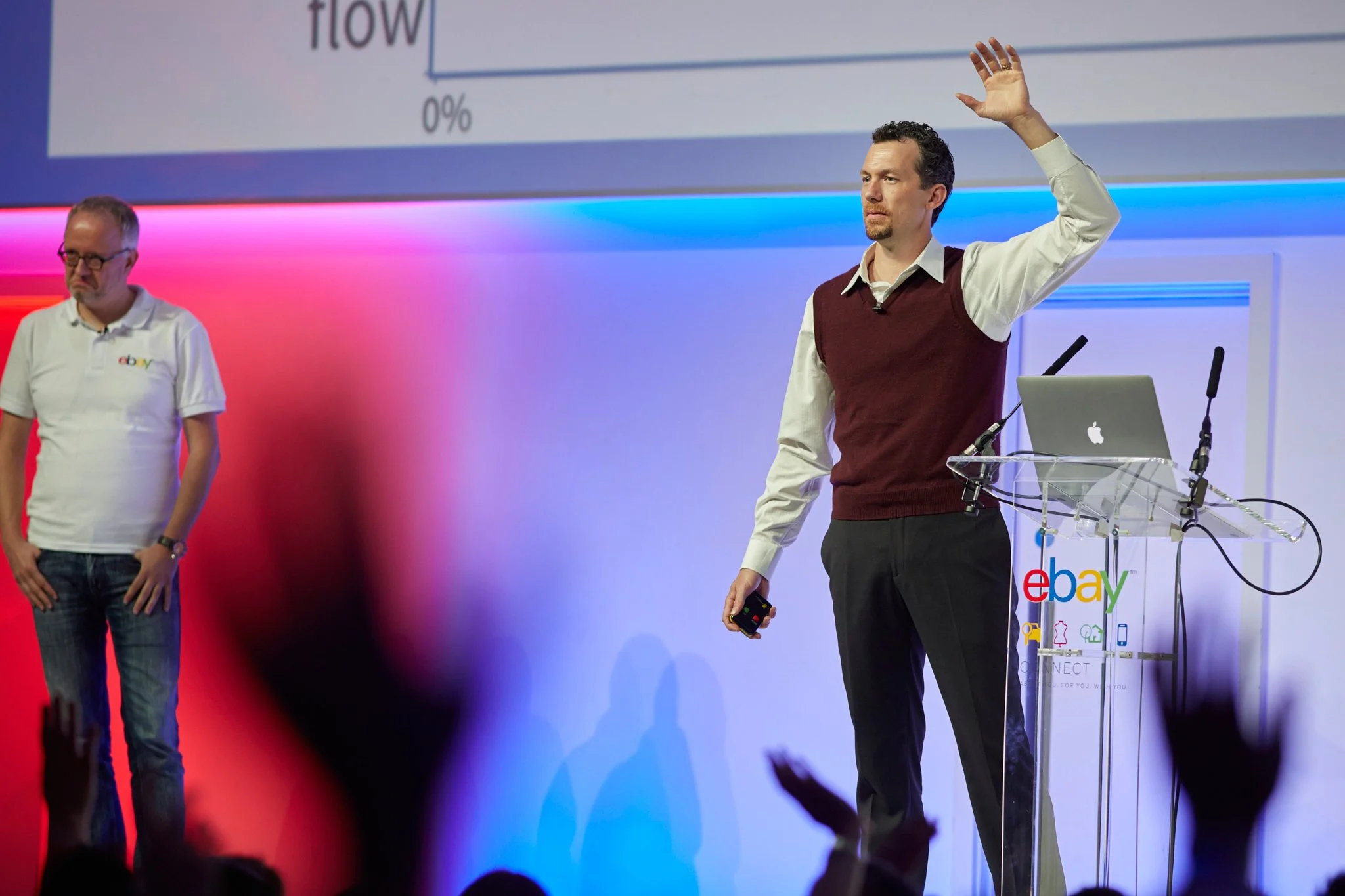 Two men on stage at an eBay presentation, one raising his hand and holding a remote, with a laptop on a clear podium, and a colorful eBay logo on a large screen behind them.