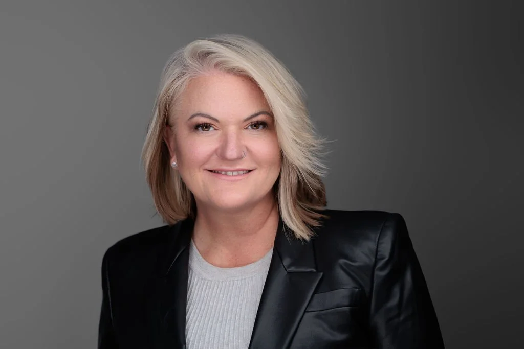 Senior Executive blonde woman with brown eyes and a wry smile wearing a black patent jacket.