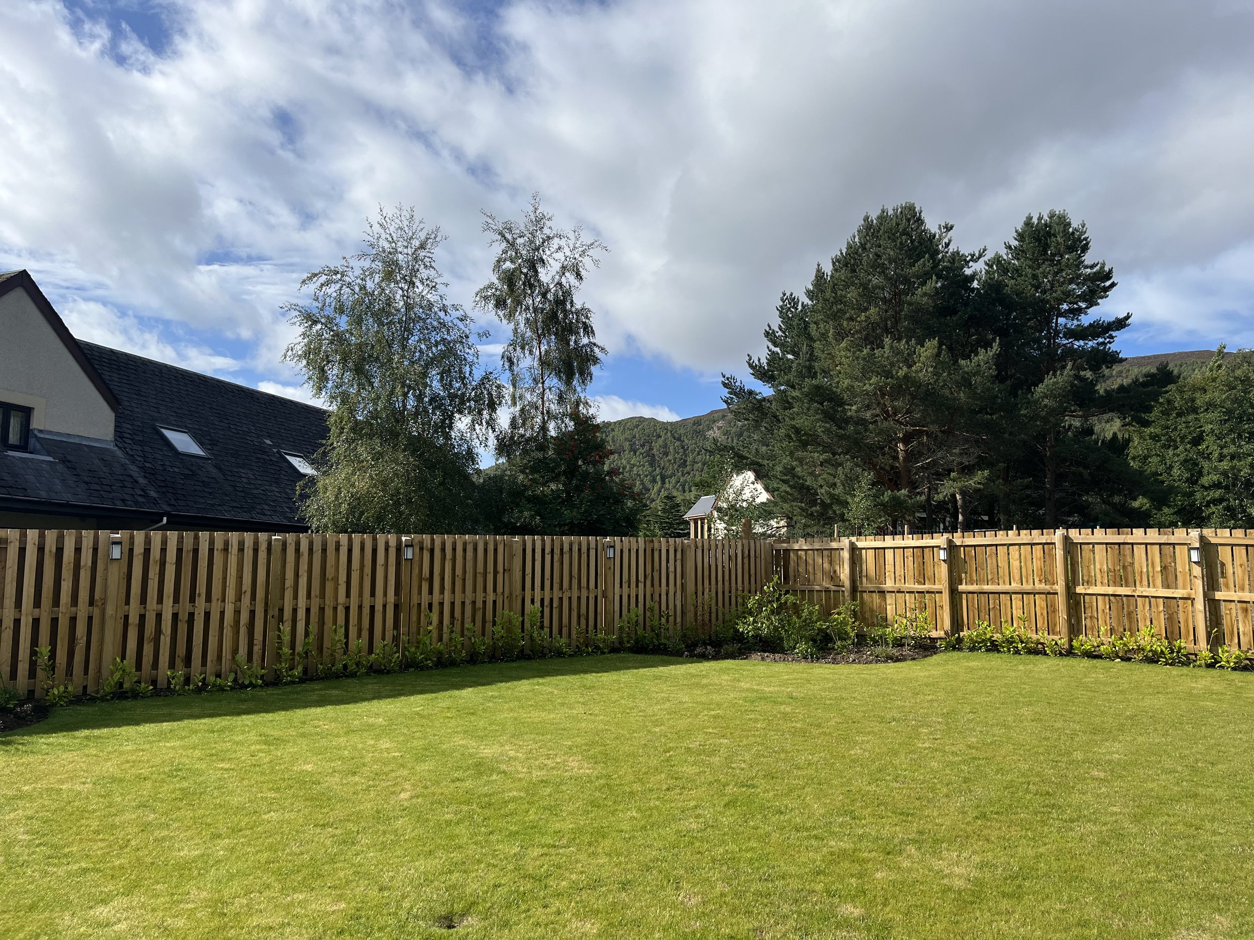 Back garden with well-maintained green lawn, wooden fence, and trees under a partly cloudy sky – private outdoor space at Aviemore holiday home.