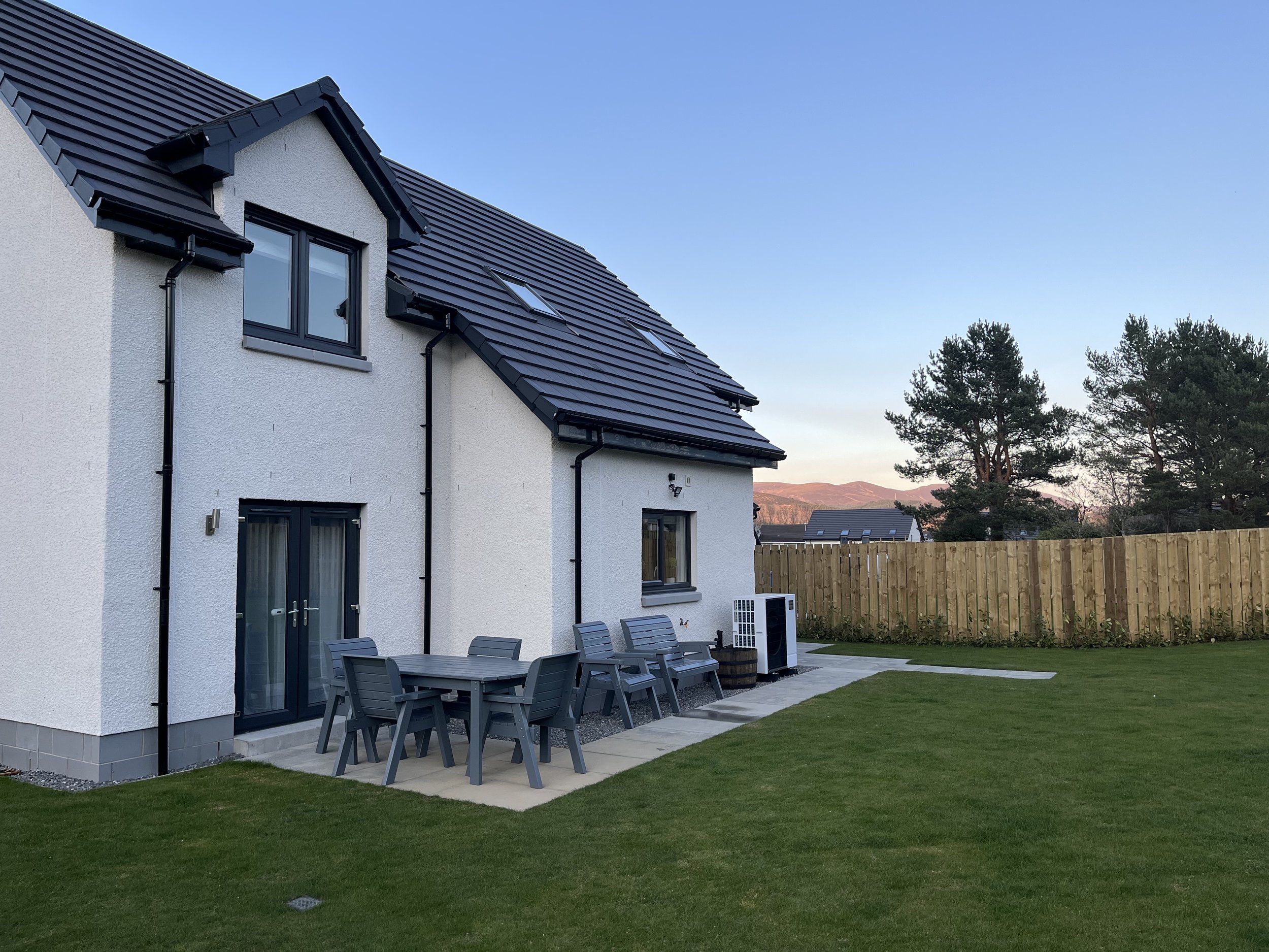 Back garden with white house, outdoor dining table and chairs, wooden fence, trees, and distant mountain at sunset – peaceful garden setting at Aviemore holiday home.