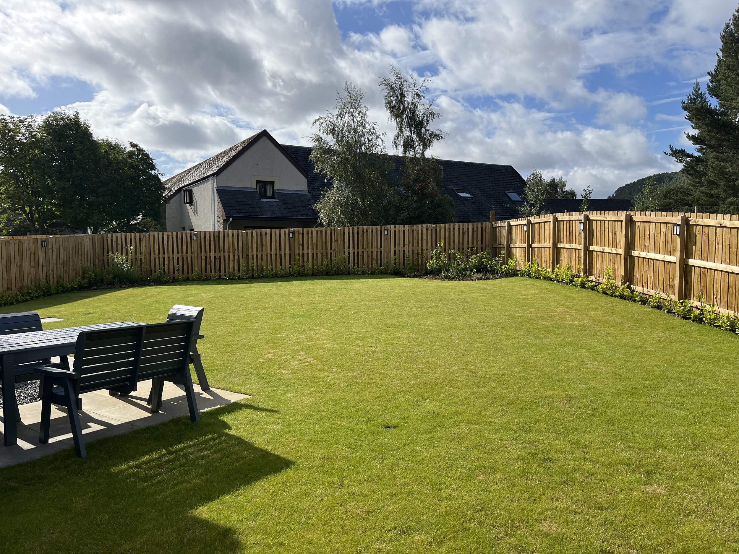 Fenced back garden with green lawn, outdoor table and chairs, bordered by plants, with houses and trees in the background under partly cloudy sky – family-friendly garden at Aviemore holiday home.