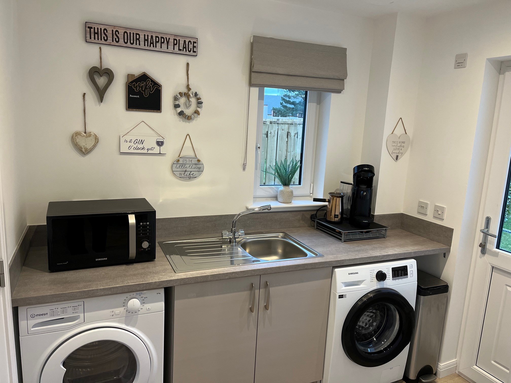 Compact laundry area with white washer and dryer beneath gray countertop, microwave, coffee machine, kettle, window with plant, and wall decor reading ‘This is our happy place’ – functional corner at Aviemore holiday home.
