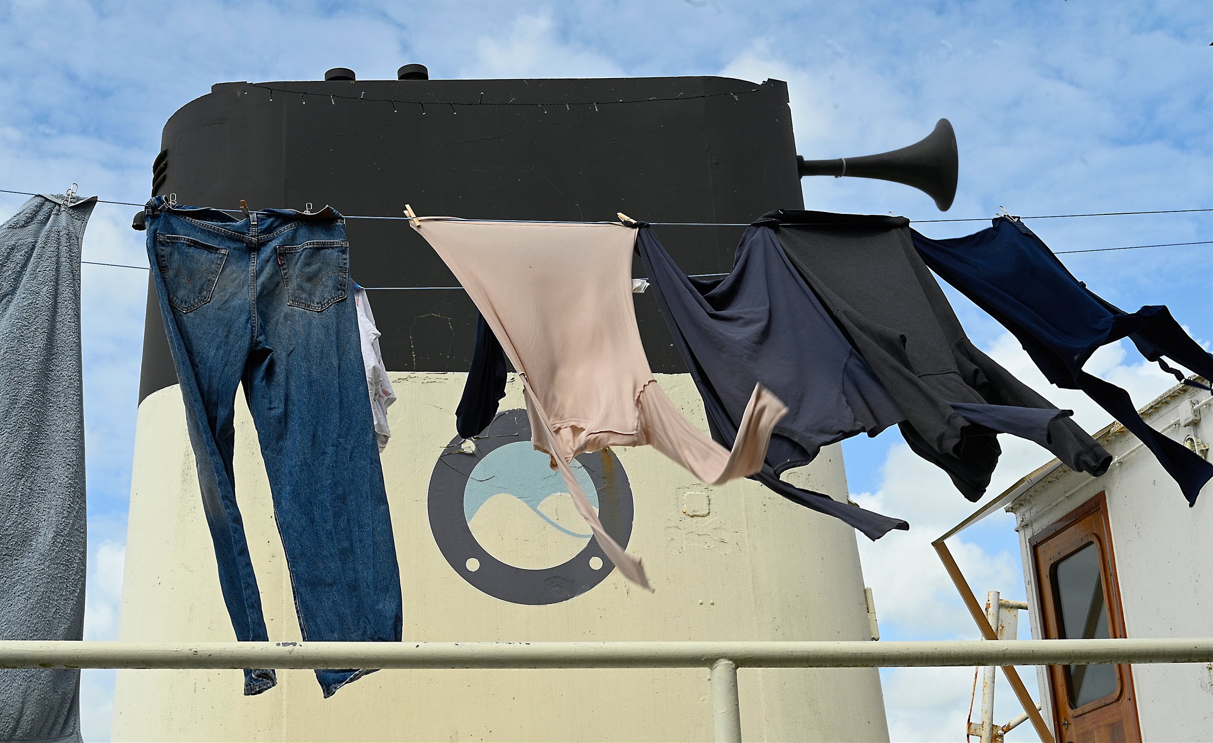Kleding hangt aan een waslijn met een zwart schip op de achtergrond en een blauwe lucht.