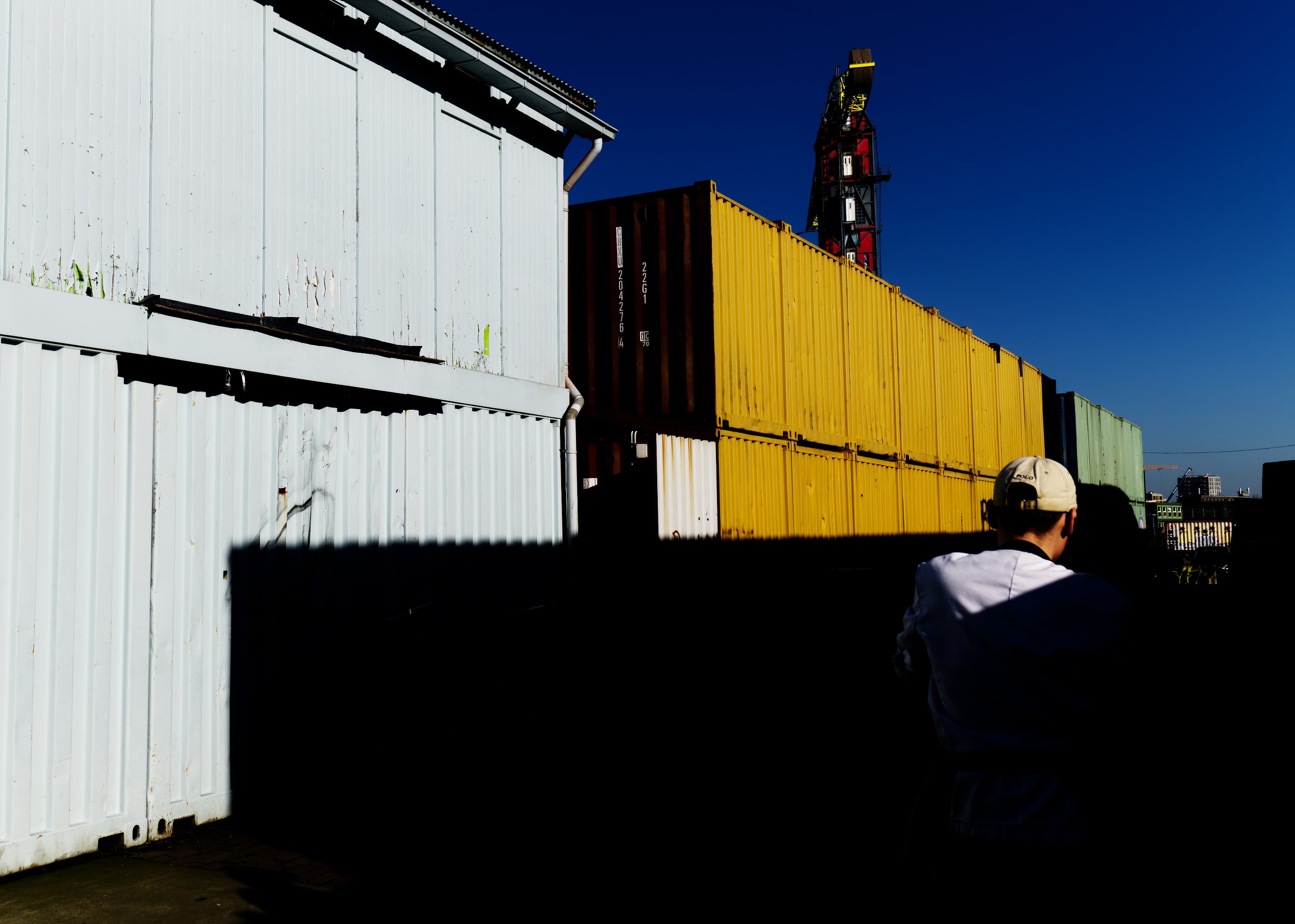 Stapel witte, gele, groene en bruine containers naast een industrieel bouwwerk, onder een heldere blauwe lucht.
