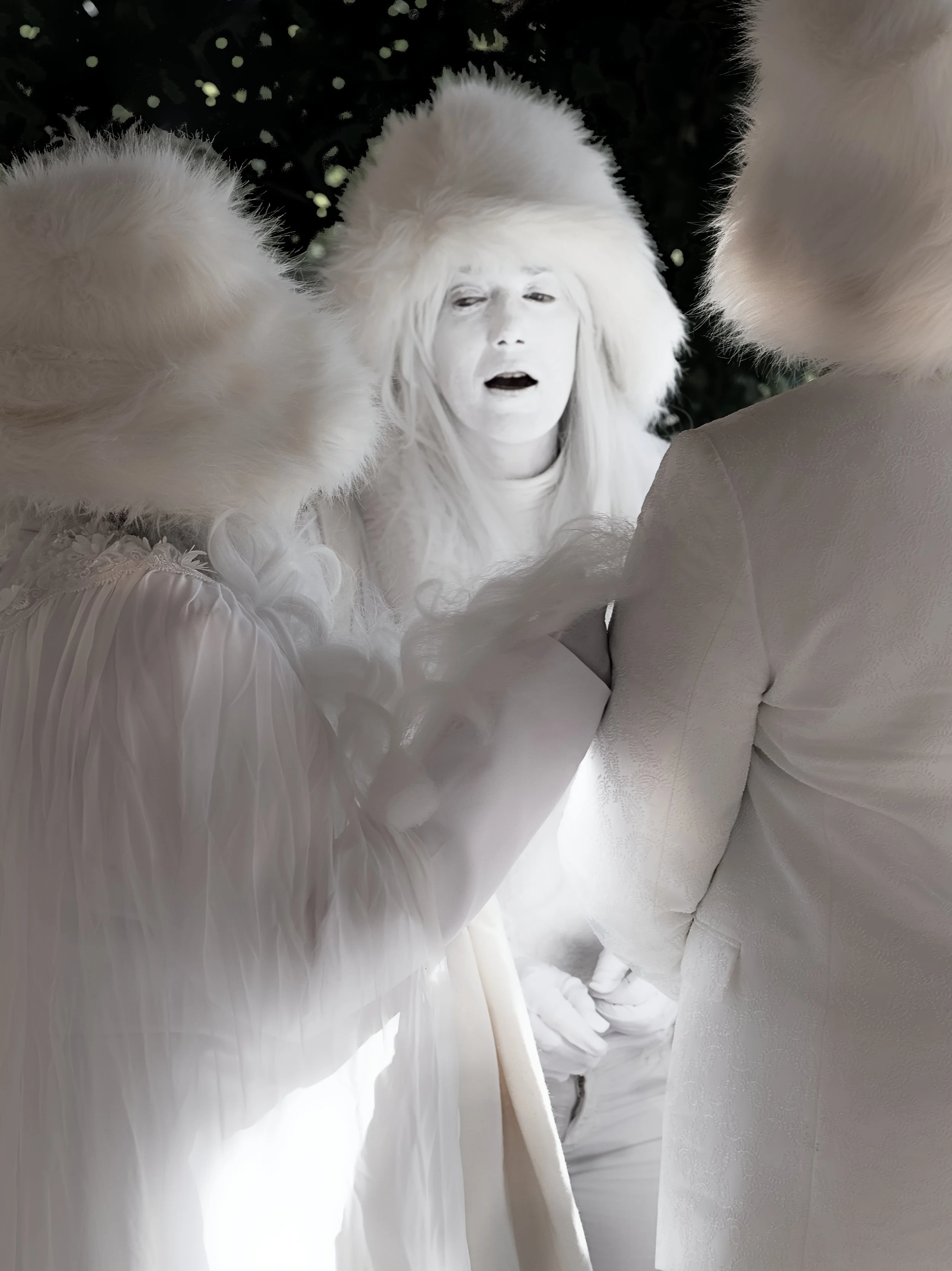 Beeld van een persoon met witte huid en wit haar, gekleed in een witte jurk met pluizige witte bontkraag, omringd door twee andere personen in witte kleding en bontkappen, op een feestelijke achtergrond.