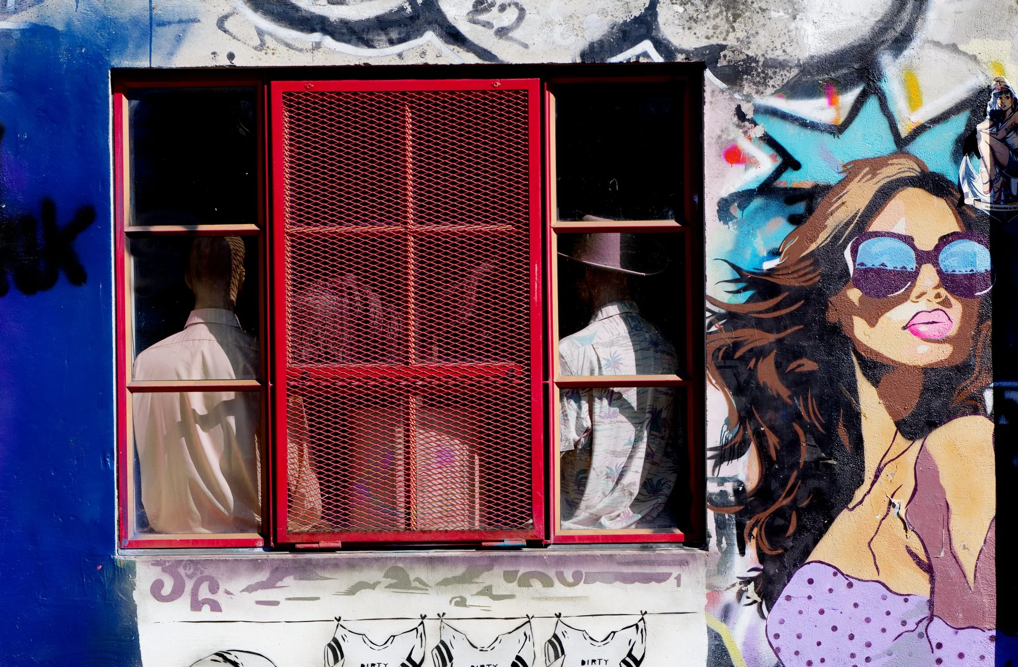 Graffiti muurschildering met een afgebeeld meisje met bruin haar, grote zonnebril en een paarse stippen top, naast een rood geverfde metalen raamwerk met drie mensen zichtbaar achter het raam.