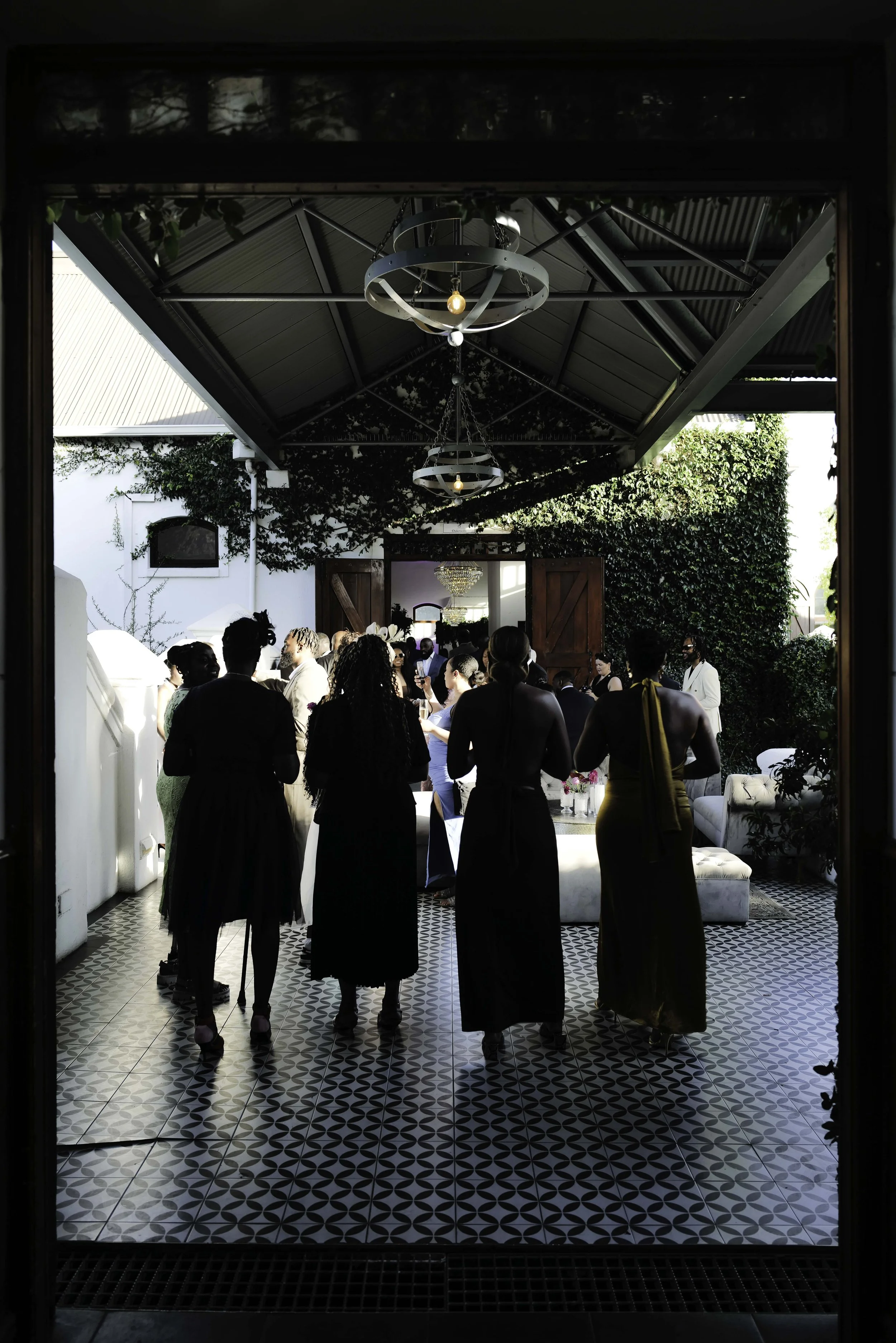 People dressed in formal attire gathered inside a bright room with tiled flooring and green ivy decor, seen through a doorway.