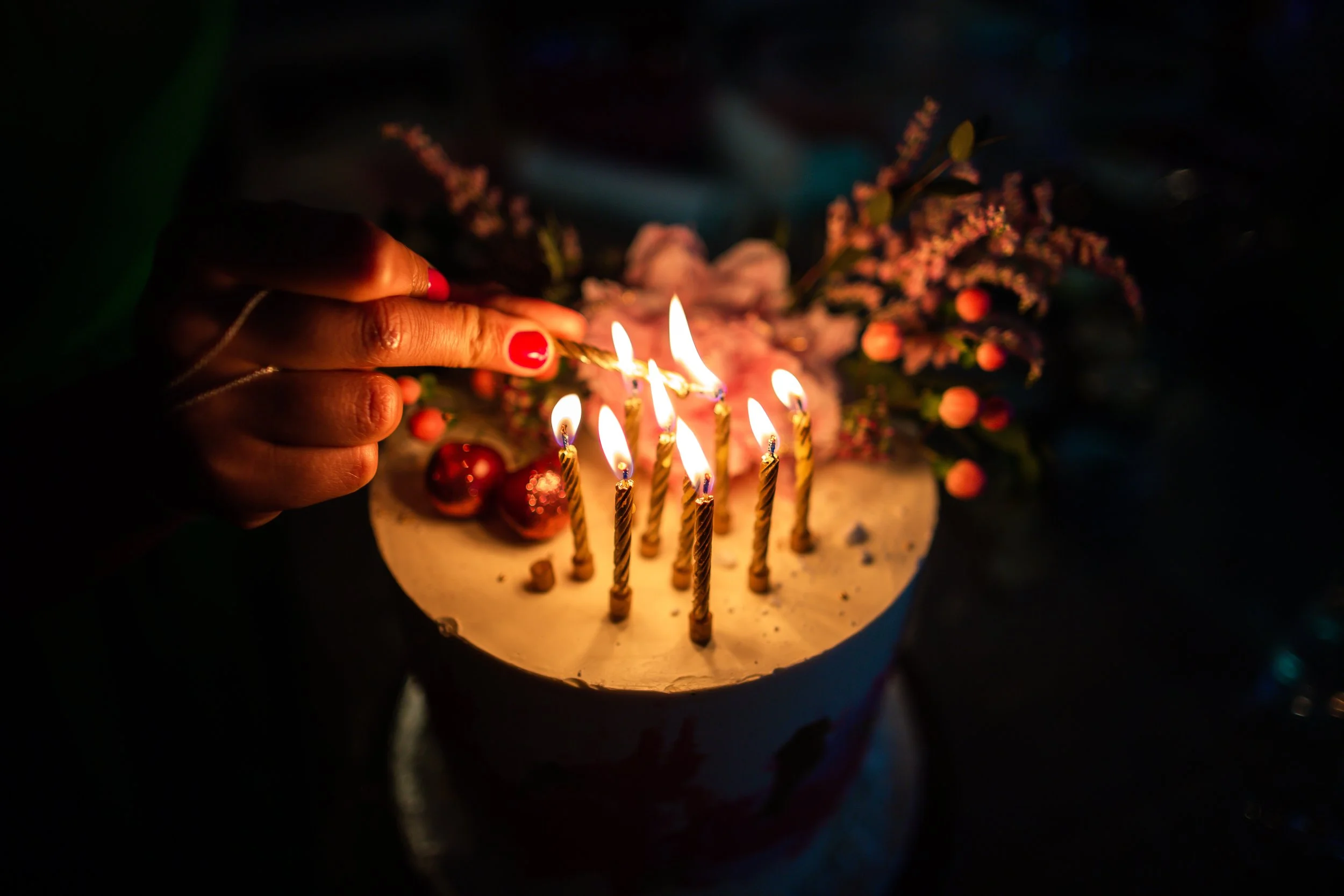 A person's hand with red painted nails lighting birthday candles on a cake decorated with flowers.