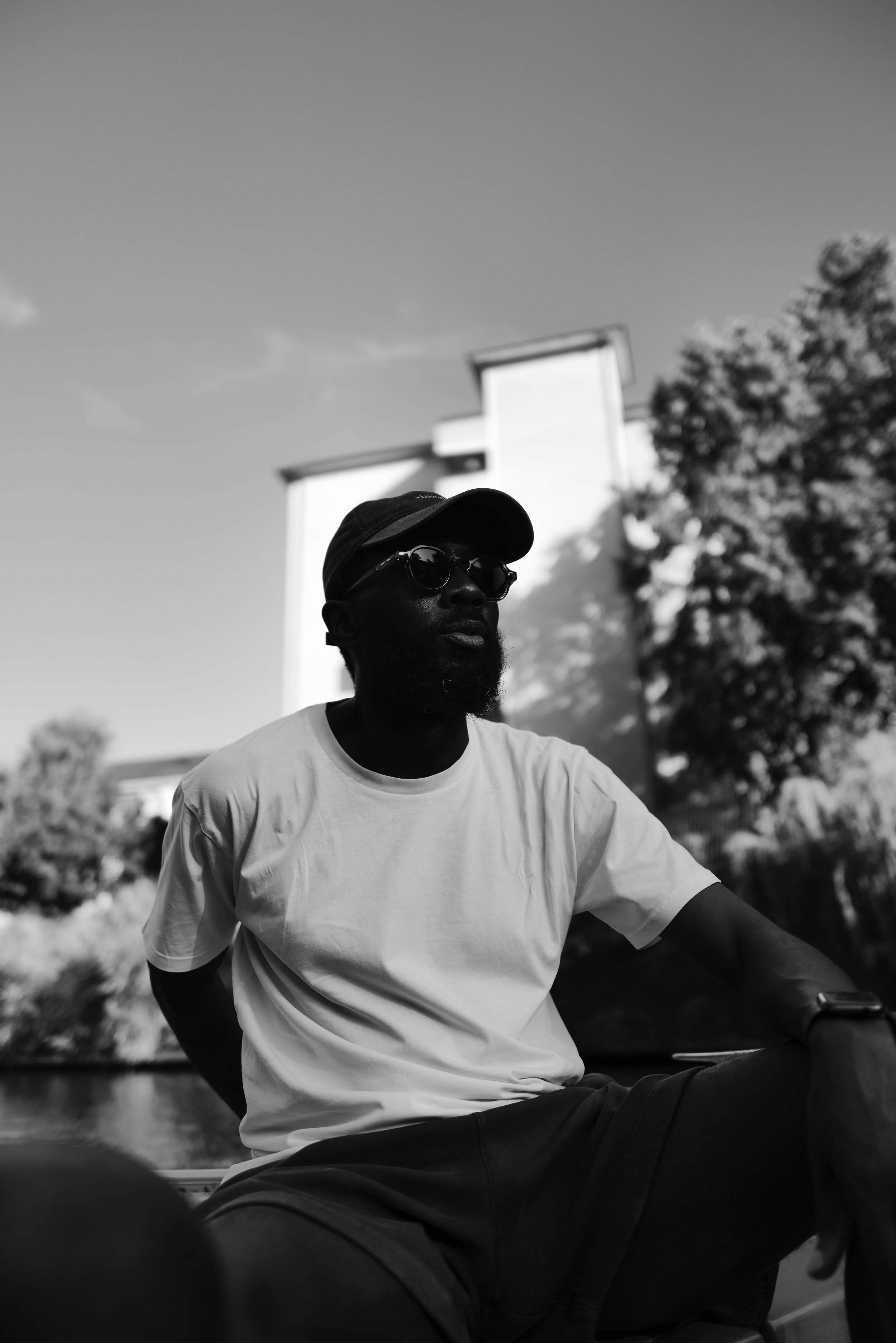A man with dark skin, wearing glasses, a cap, and a white t-shirt, sitting outdoors with trees and a building in the background, in black and white.