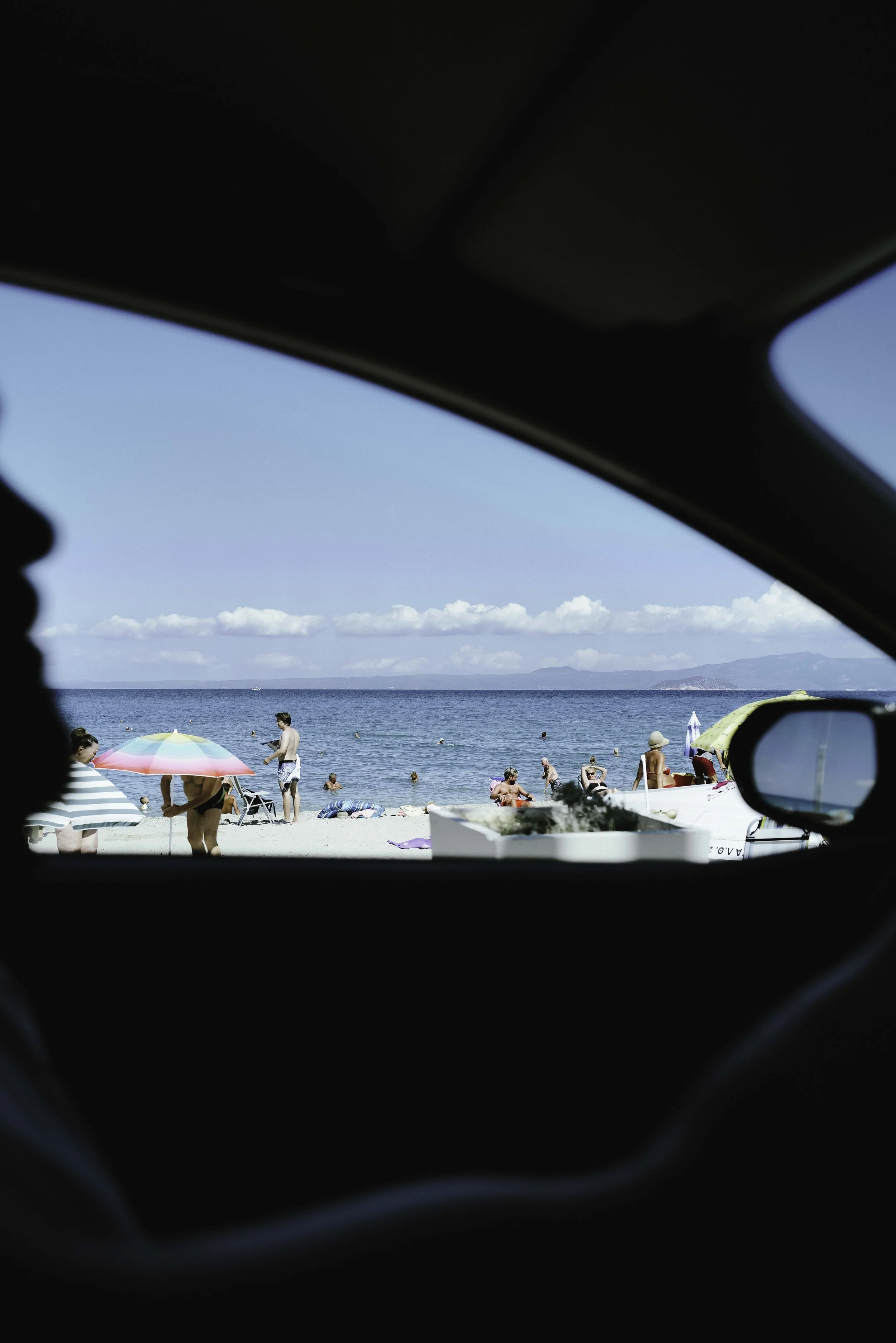 Beach scene viewed from inside a vehicle, showing people relaxing with umbrellas, swimming, and enjoying the sunny weather by the shore with mountains in the distance and a blue sky with clouds.
