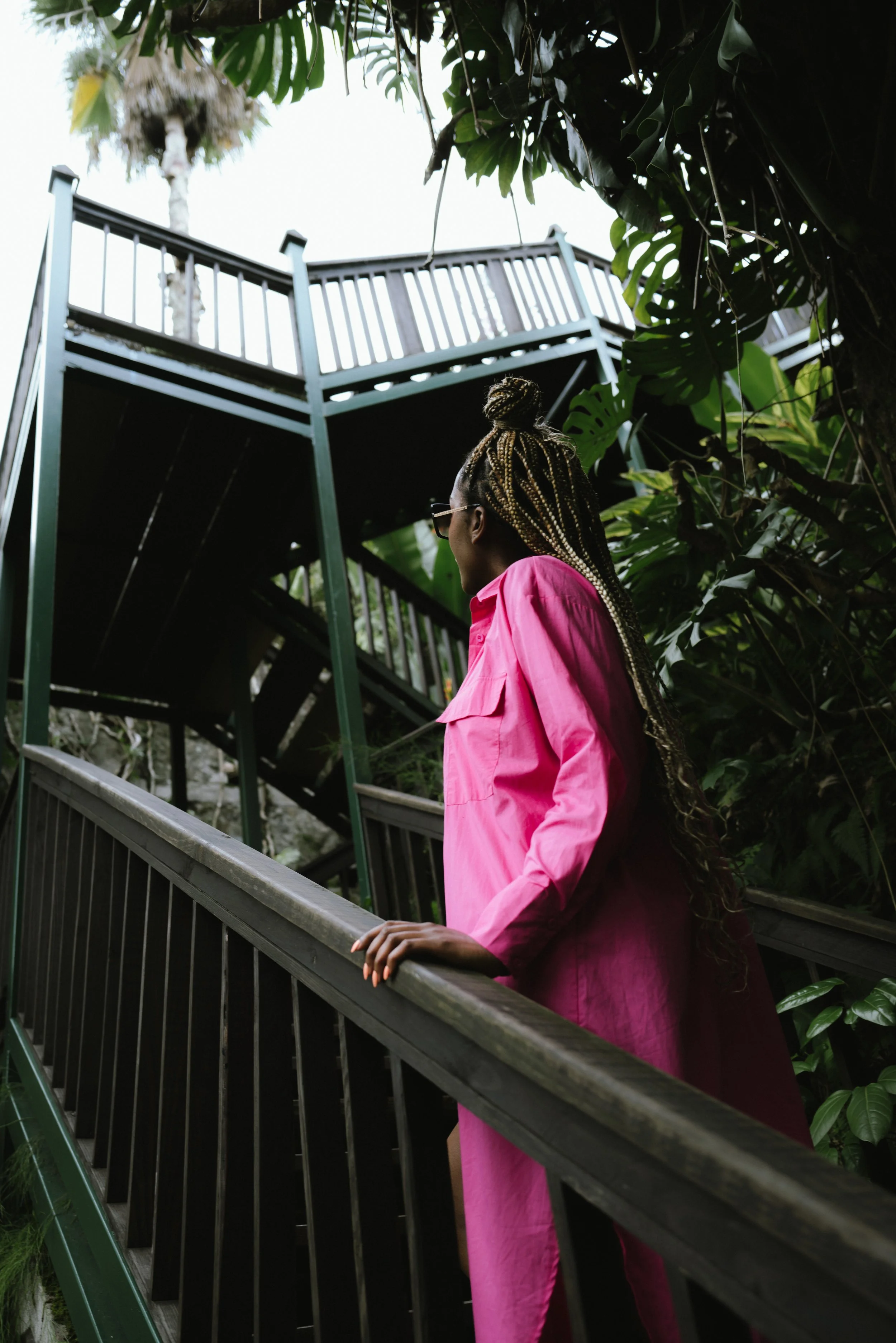 A woman with braided hair and glasses, wearing a pink jacket, stands on a staircase surrounded by lush green plants, looking up at a tall outdoor balcony structure.