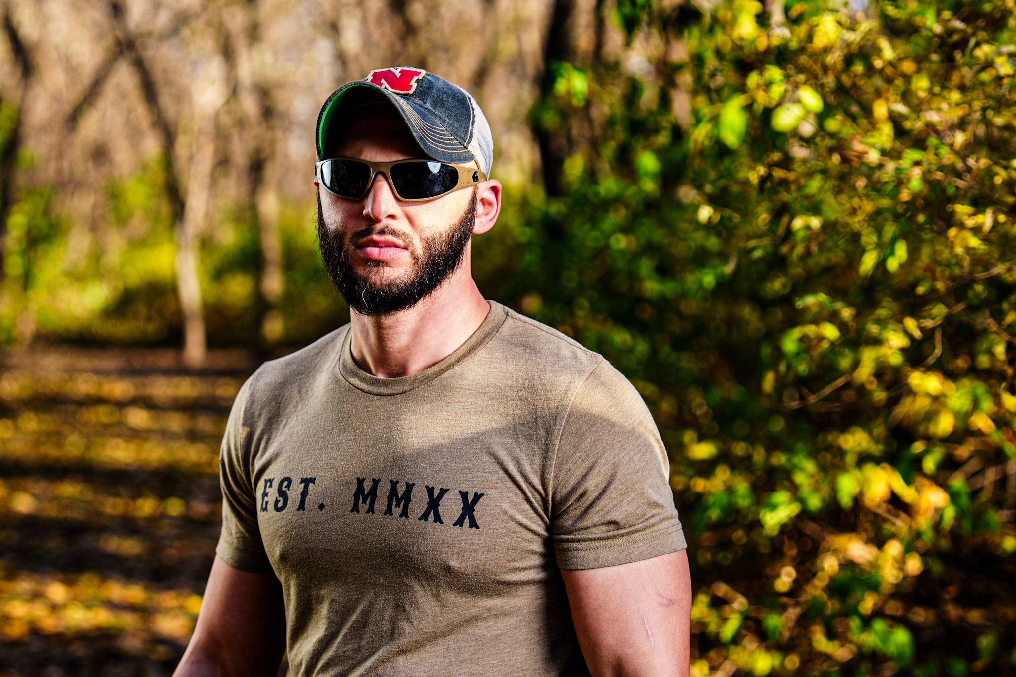 A man with a beard wearing sunglasses, a baseball cap with an 'N' on it, and a beige t-shirt in a park with autumn foliage.