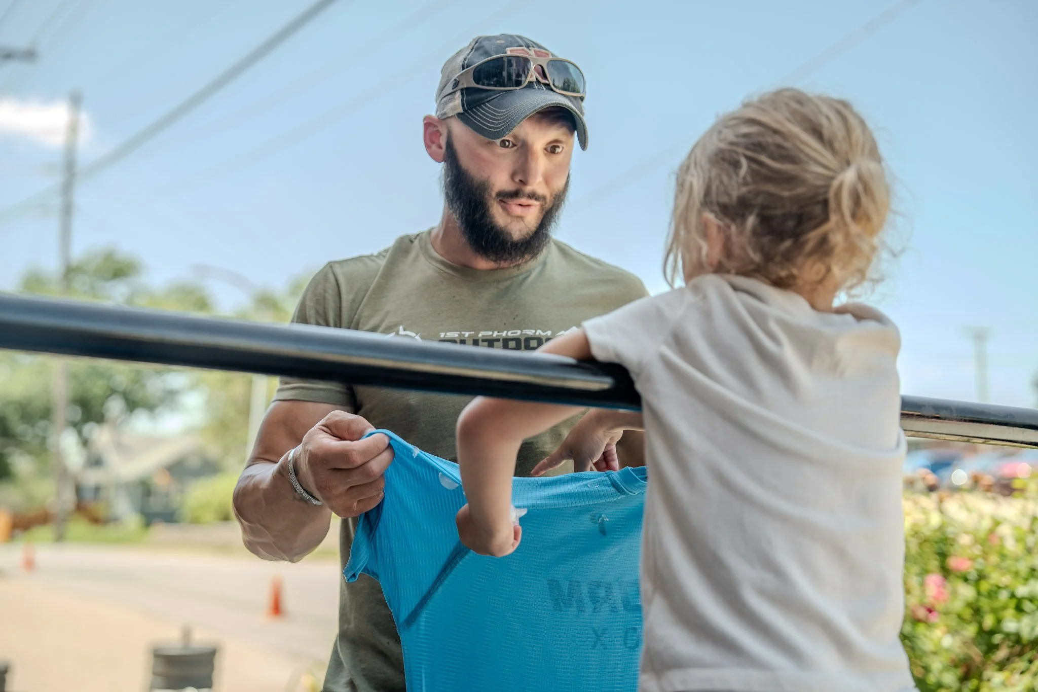A young man with a beard, wearing sunglasses on his cap, is showing a bright blue shirt to a young girl from the other side of a metal fence at an outdoor event.