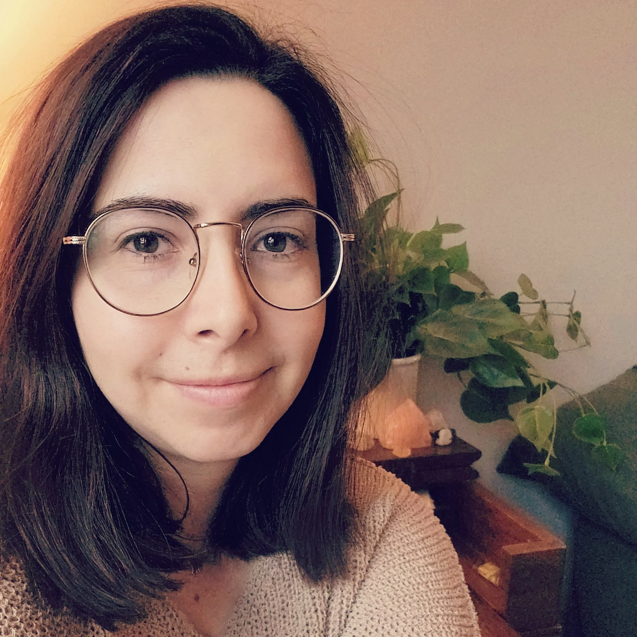 A woman with shoulder-length brown hair and glasses smiling indoors near a potted plant and a lamp.