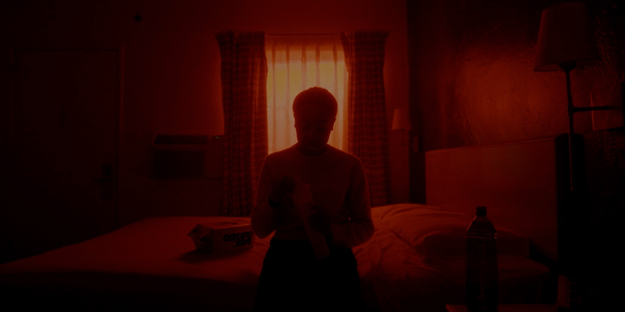Silhouette of a person standing in a dimly lit room with orange light coming from a window with checkered curtains, a bed, a water bottle on a side table, and a box of snacks.