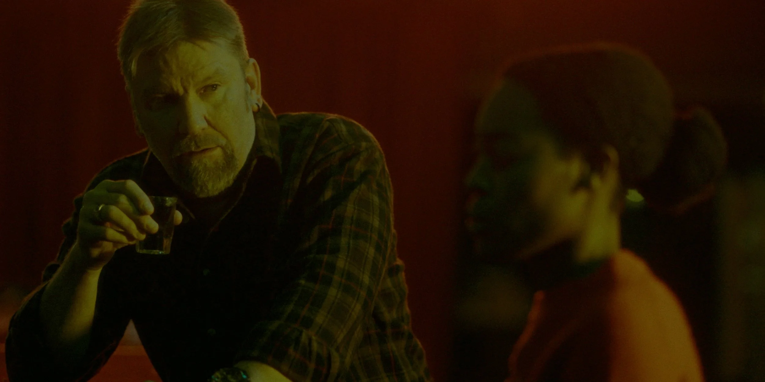 A man with gray hair and beard holding a shot glass, sitting in a dimly lit bar with a reddish background, engaged in conversation with a woman with braided hair and glasses.