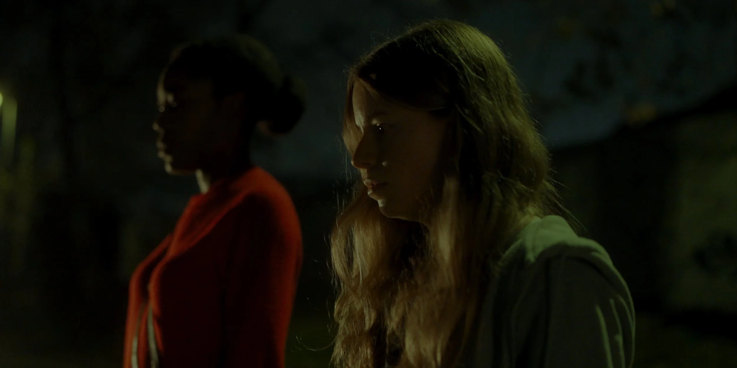 Two women standing outdoors at night, both with serious expressions, one with dark hair in a bun wearing a red coat, the other with long blonde hair wearing a light-colored hoodie.